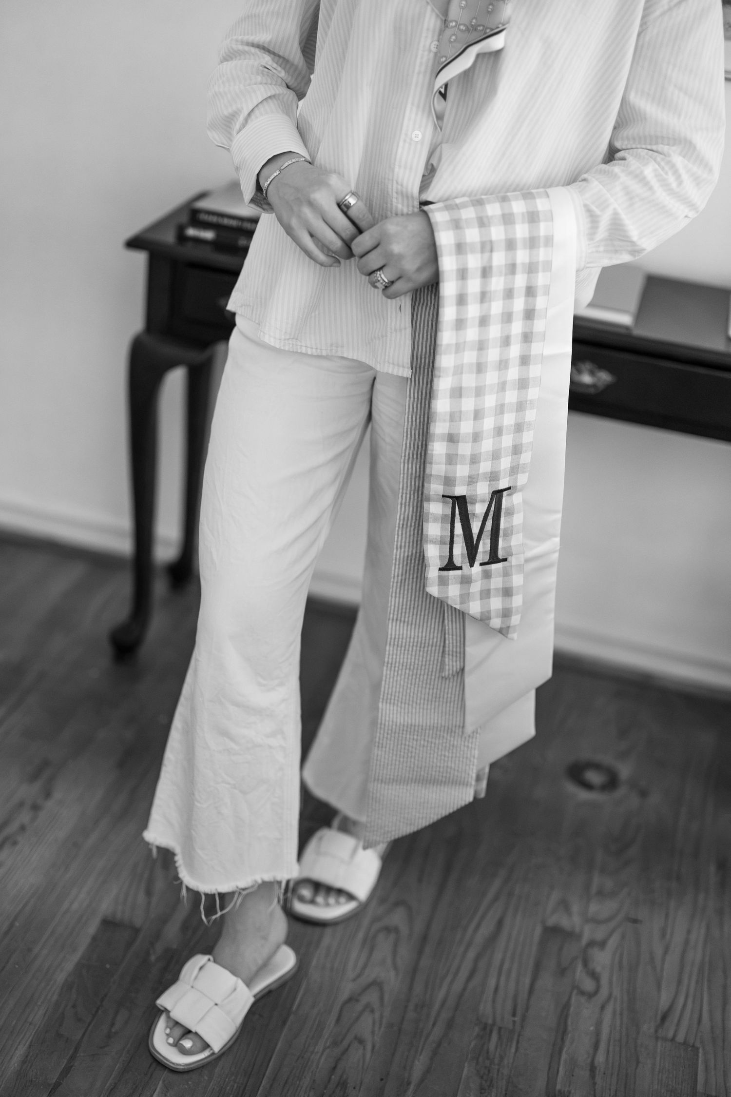 Person wearing white shirt, cropped jeans, and slide sandals, holding a gingham monogrammed towel with letter M.
