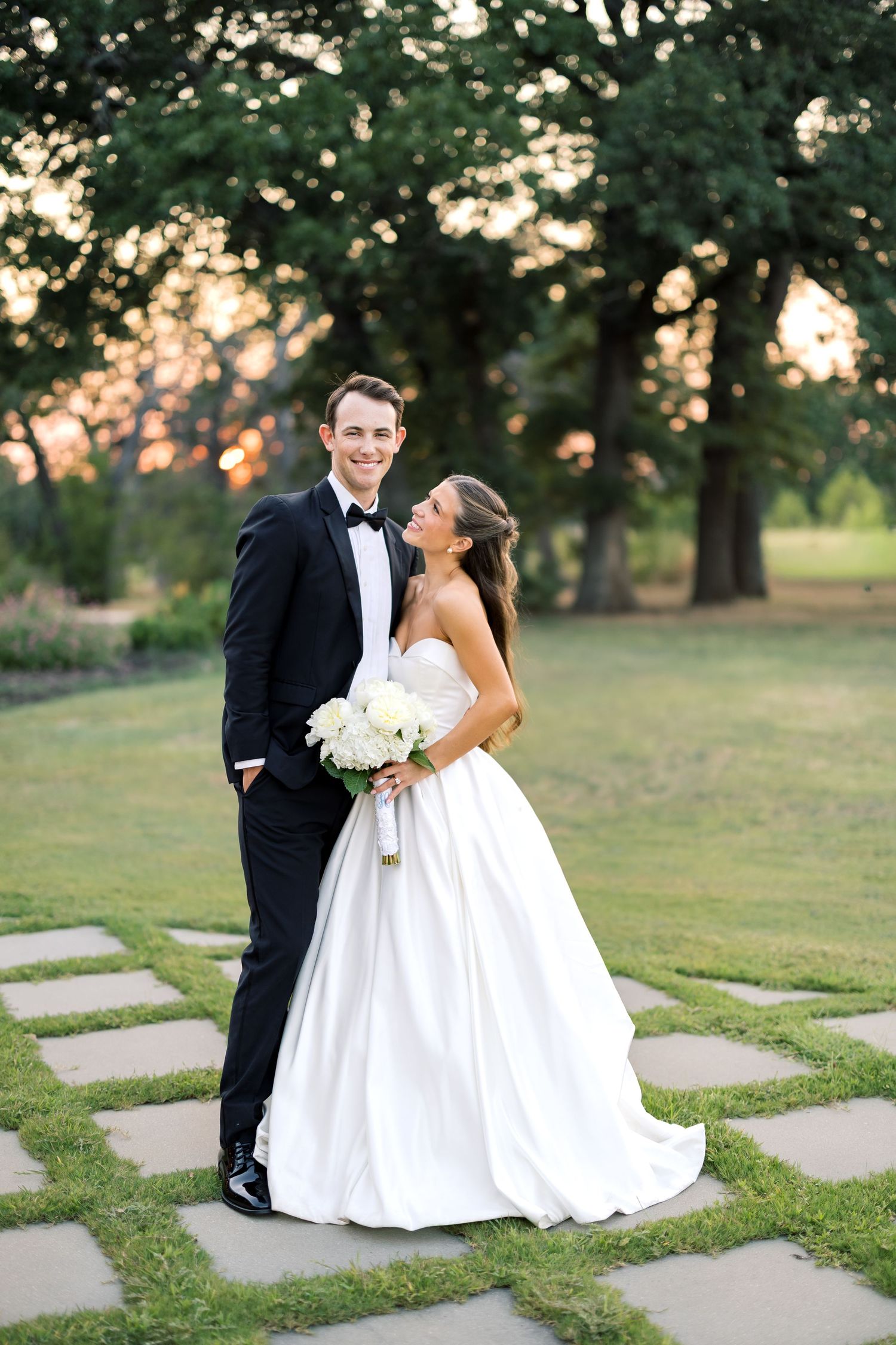 A bride and groom pose elegantly at sunset, she in a strapless ballgown holding white flowers, he in a classic black tuxedo.