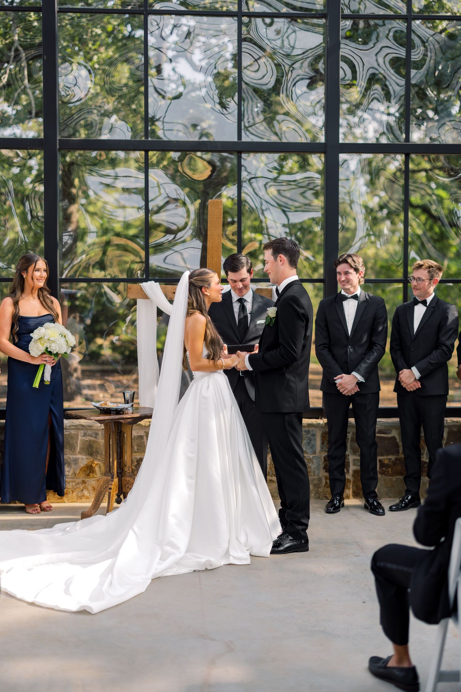 Bride and groom exchange vows at elegant indoor wedding ceremony with groomsmen in black tuxedos and large glass windows.