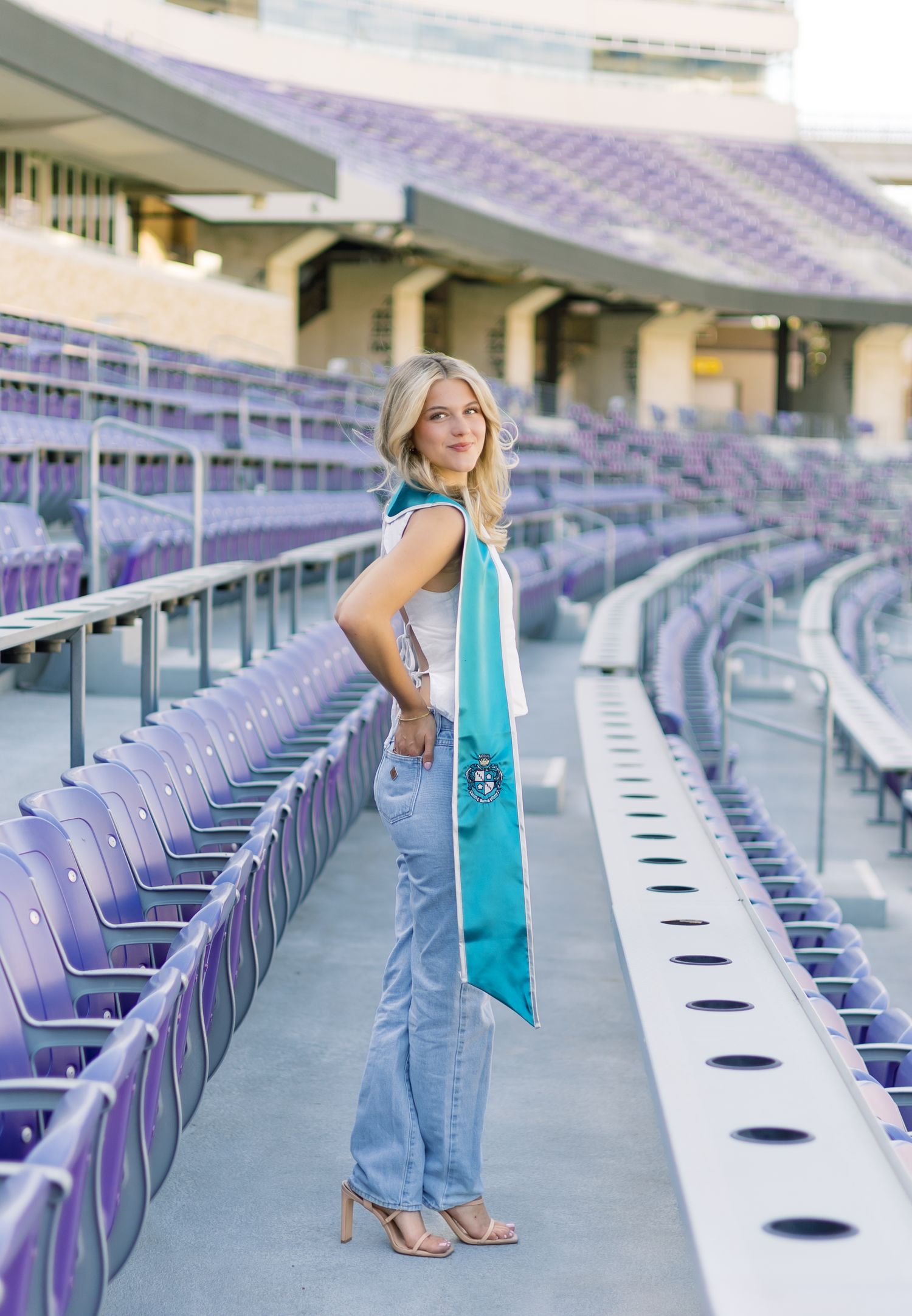 Blonde woman in jeans and white top wearing teal ZTA graduation stole in purple stadium bleachers.