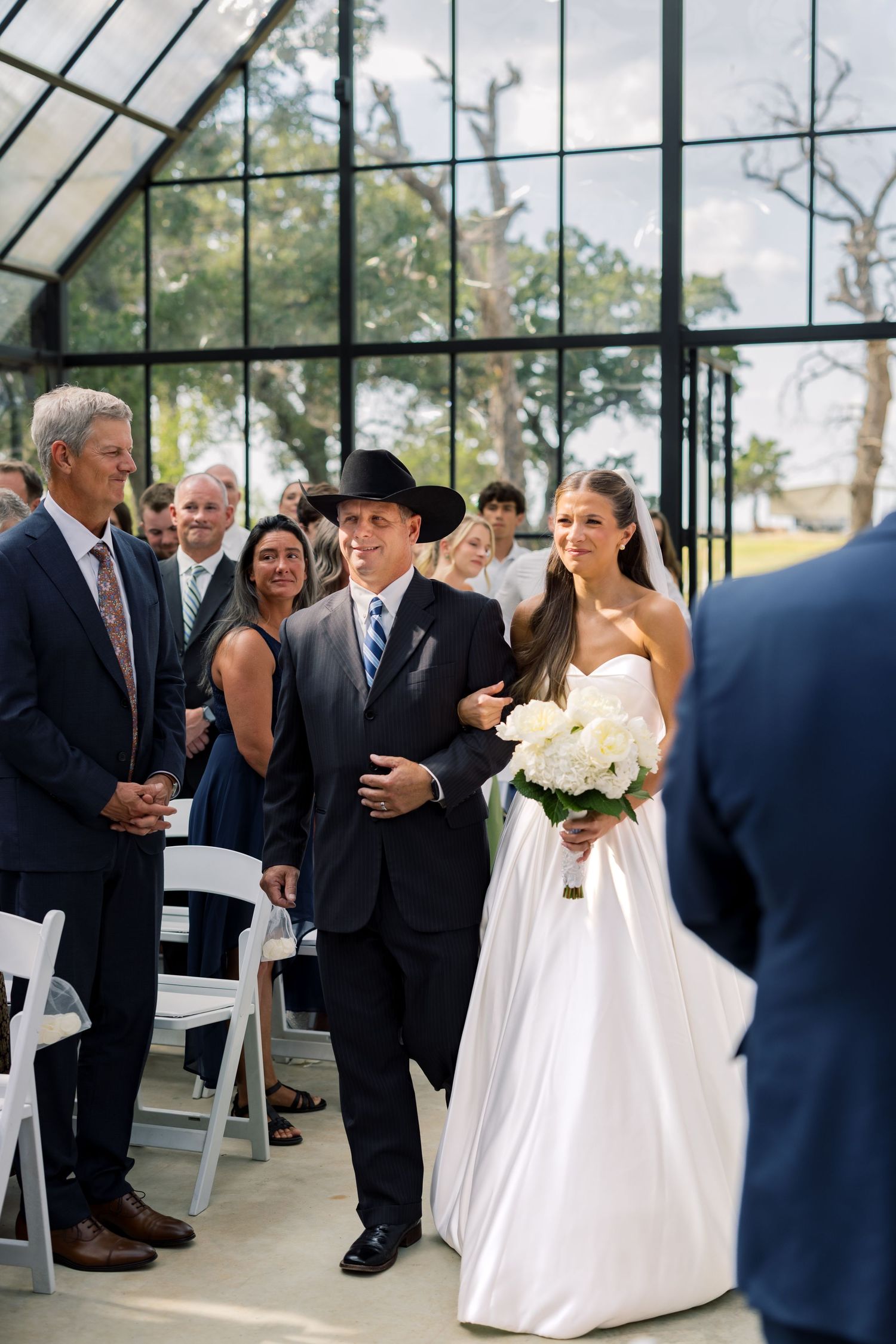 Bride in white gown walks down aisle with escort in black cowboy hat during outdoor wedding ceremony.