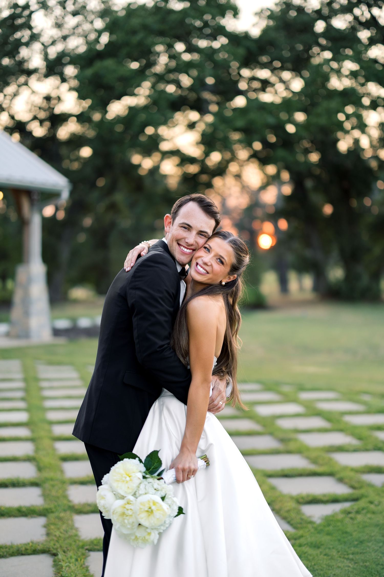 Smiling bride and groom embrace outdoors at sunset, bride holding white peony bouquet in elegant white gown.