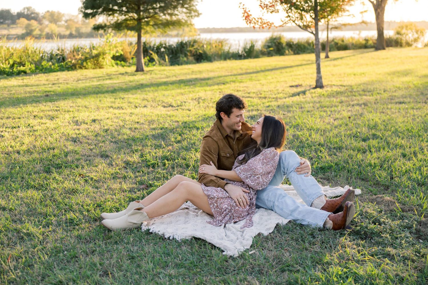 Couple sitting on a blanket in a grassy park near water, leaning in close during sunset.