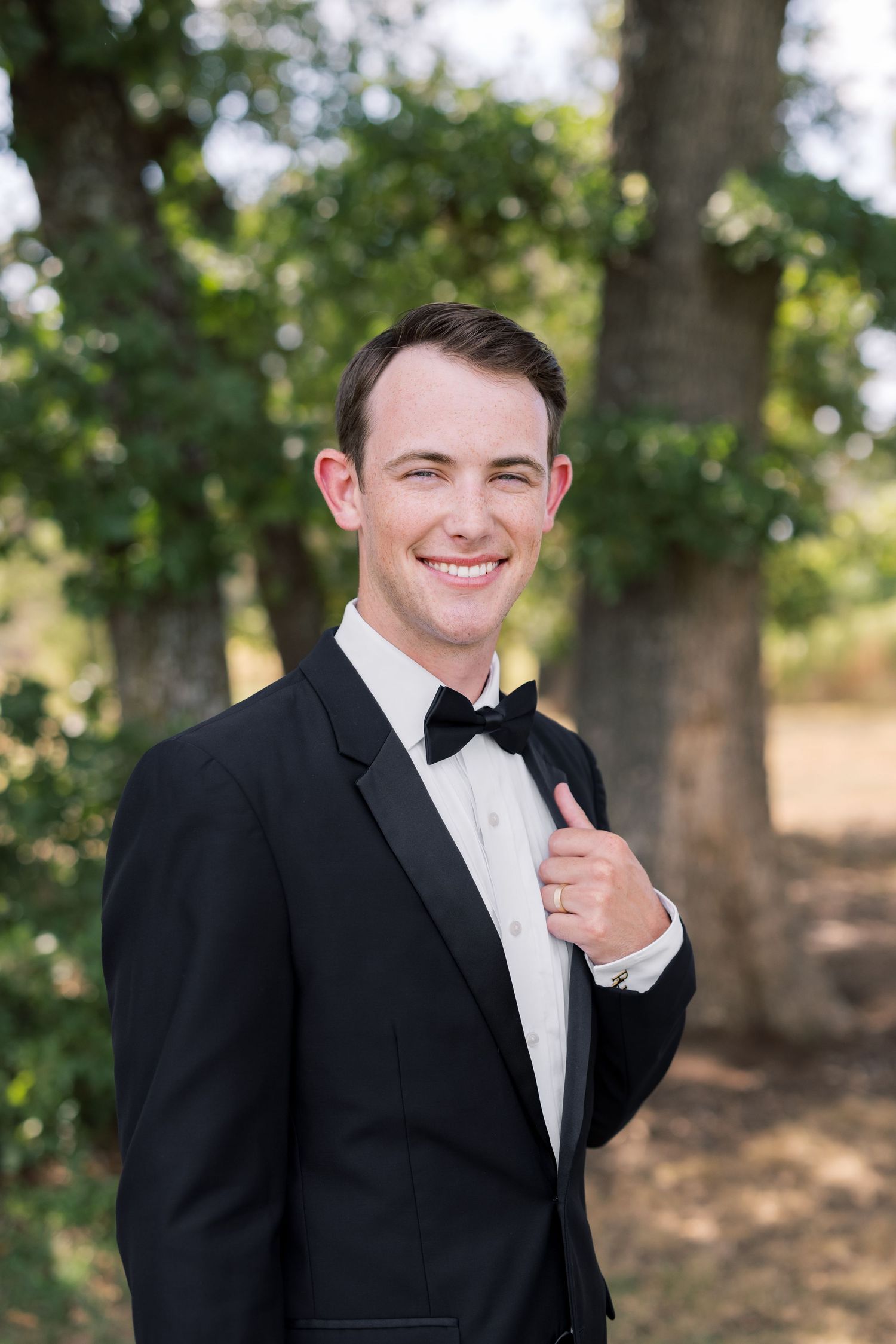 Smiling young man in black tuxedo and bow tie posing outdoors with lush green trees in the background.