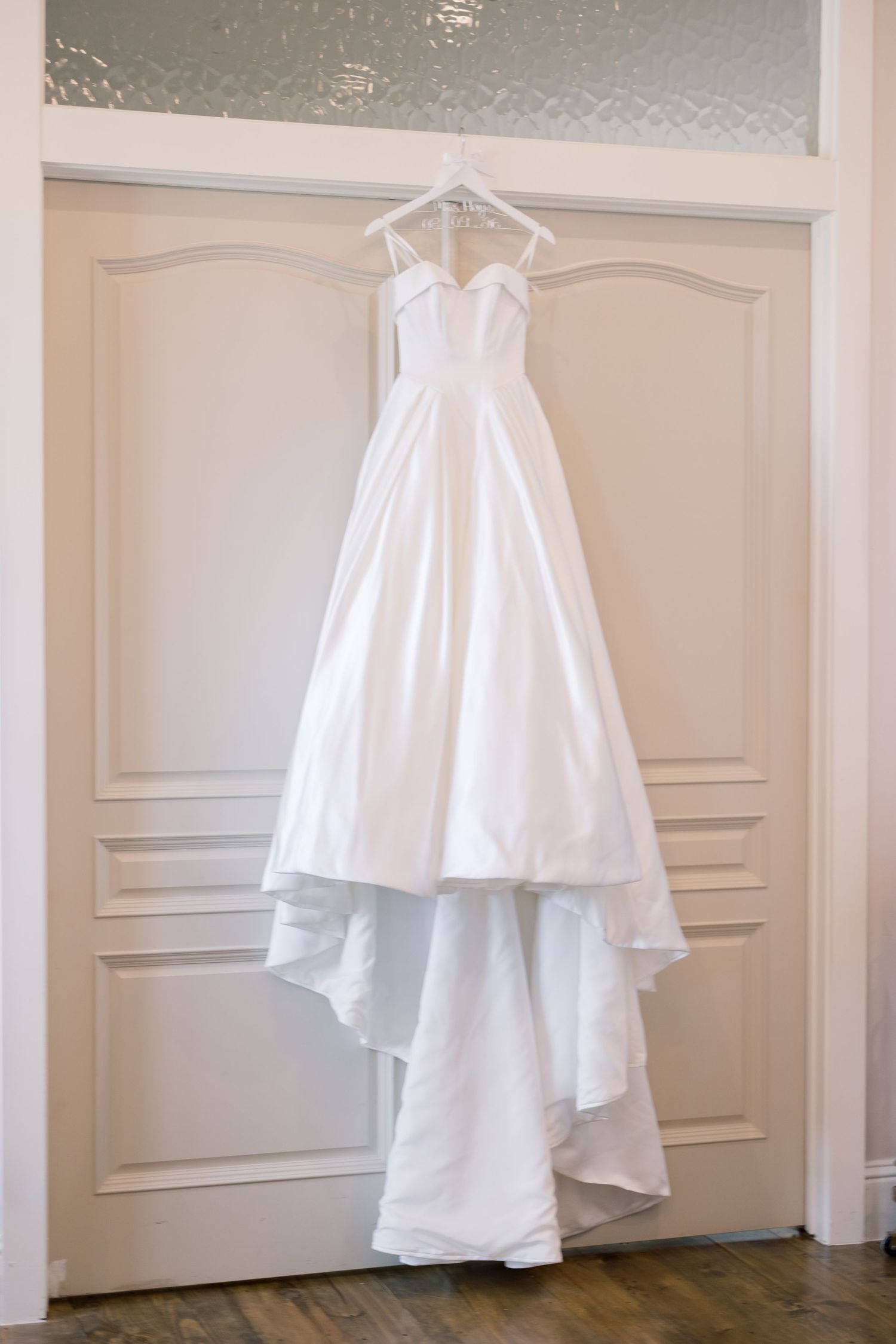 Elegant white strapless ball gown wedding dress hanging on a door hanger in a bright, neutral-toned bridal suite.
