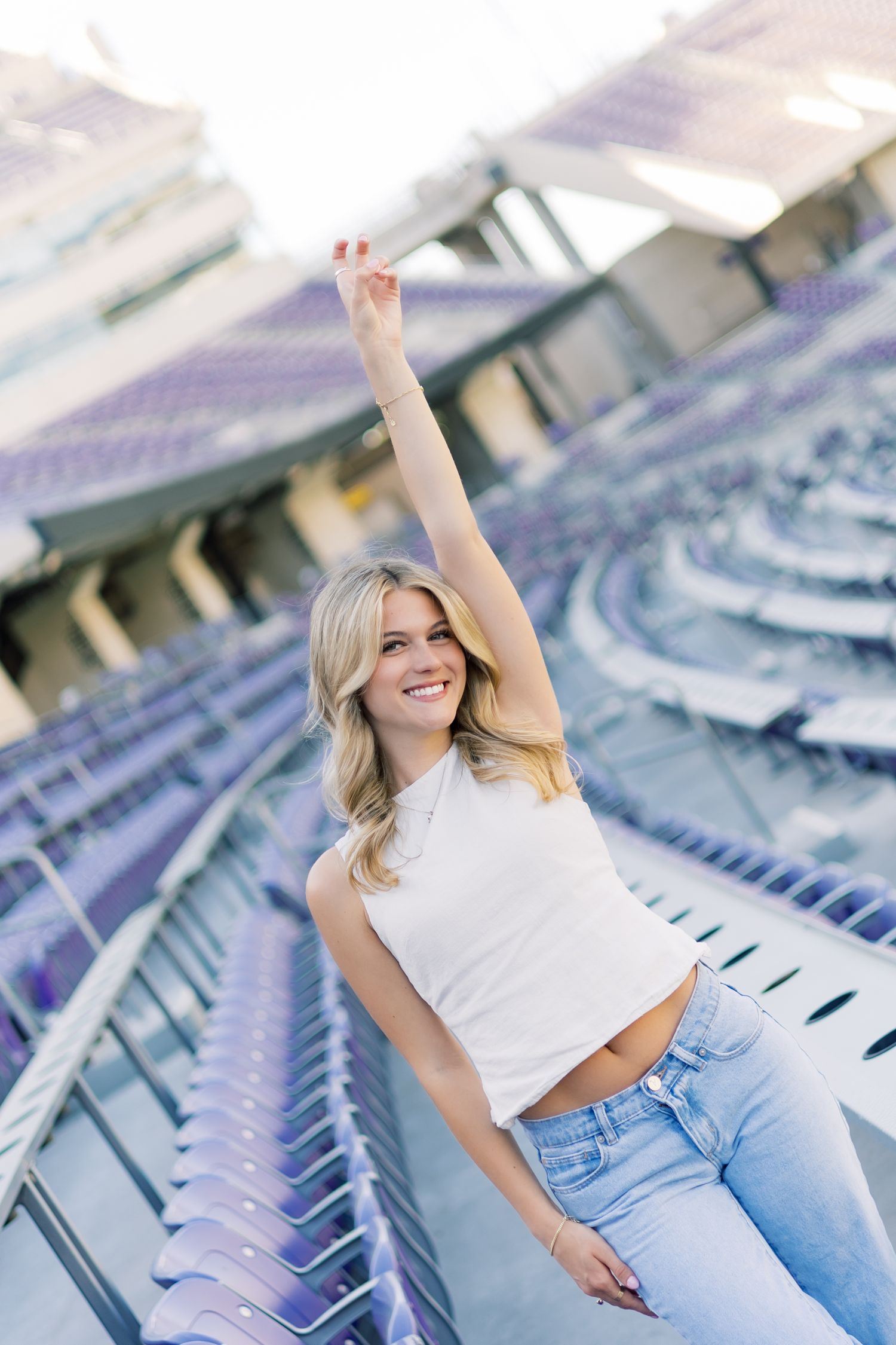 Joyful blonde woman in white crop top and jeans raising arm in empty purple stadium.