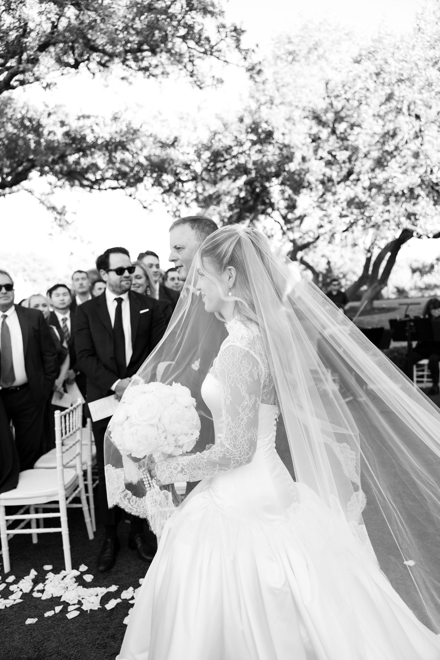 Black and white photo of bride in ballgown and veil walking down petal-lined wedding aisle.
