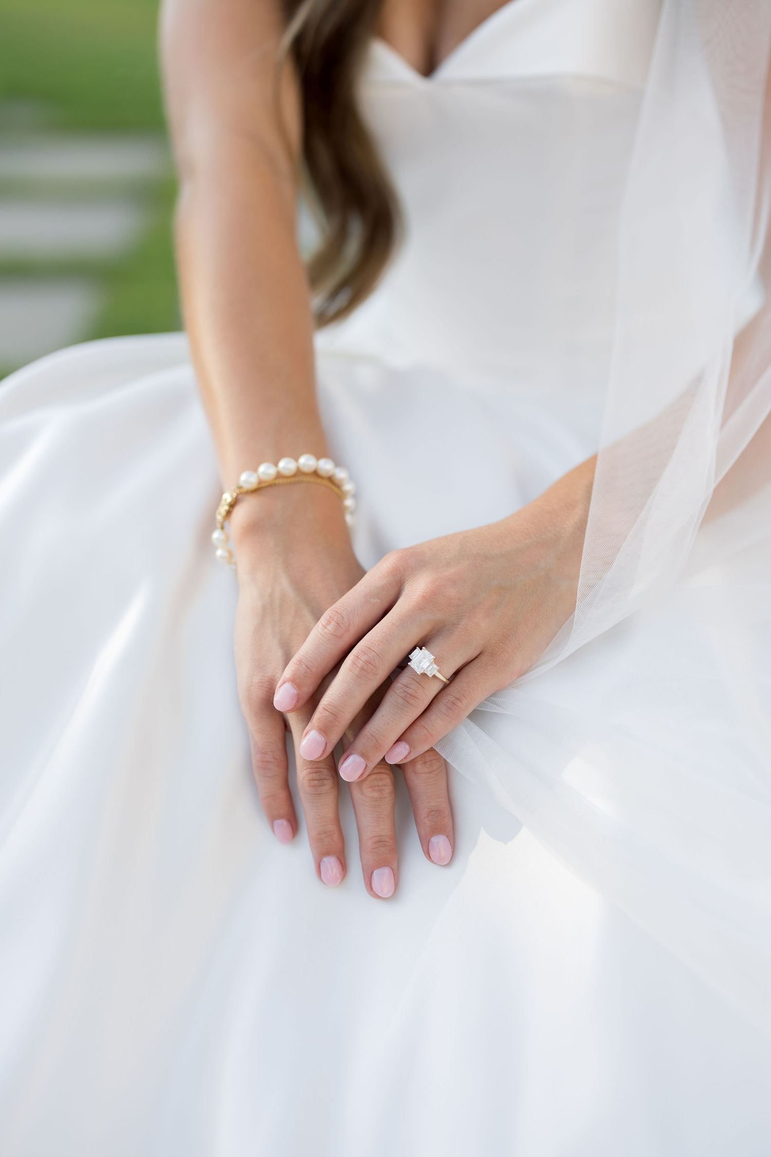 Bride's hands resting on white wedding dress, wearing diamond engagement ring and pearl bracelet with pink manicure.
