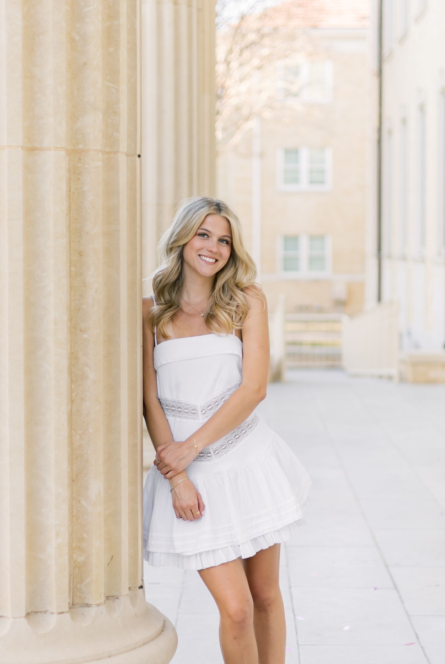Smiling blonde woman in white strapless dress leaning against a stone column on a sunny campus.