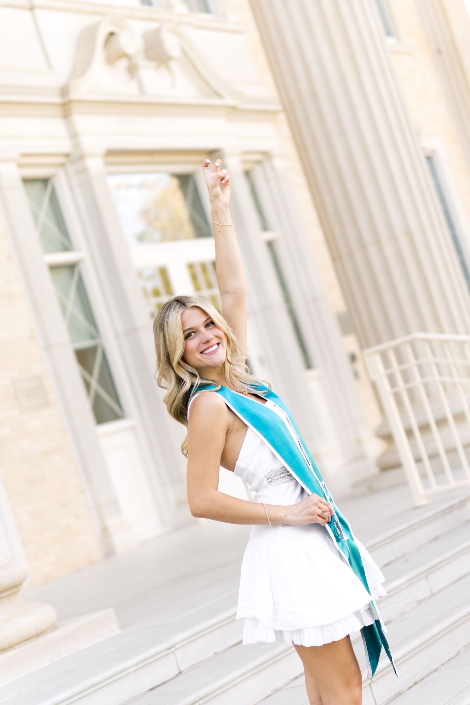 Joyful blonde graduate in white dress and teal sash raising arm in celebration outside university.