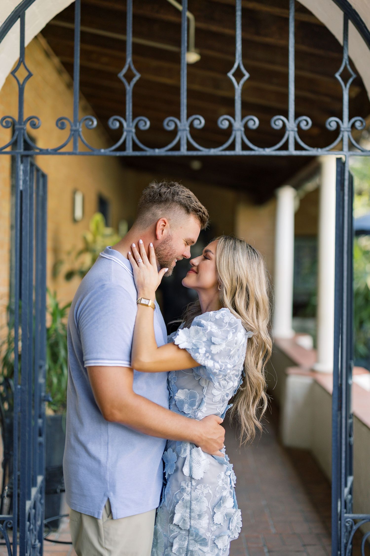 Couple sharing a romantic embrace and almost kissing under an ornate iron gate archway outdoors.