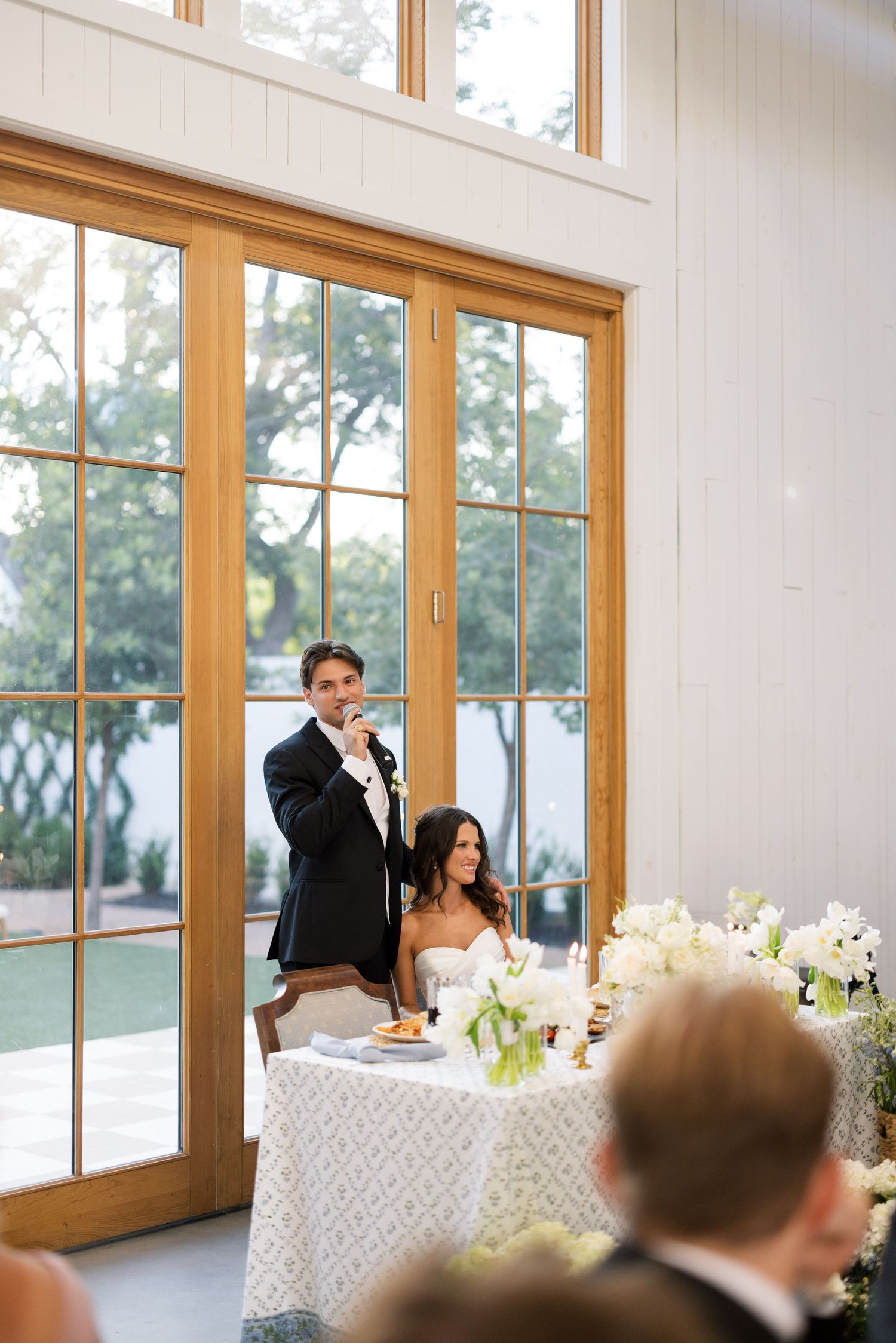 Groom giving a wedding speech at reception while bride sits at sweetheart table, elegant white floral decor.