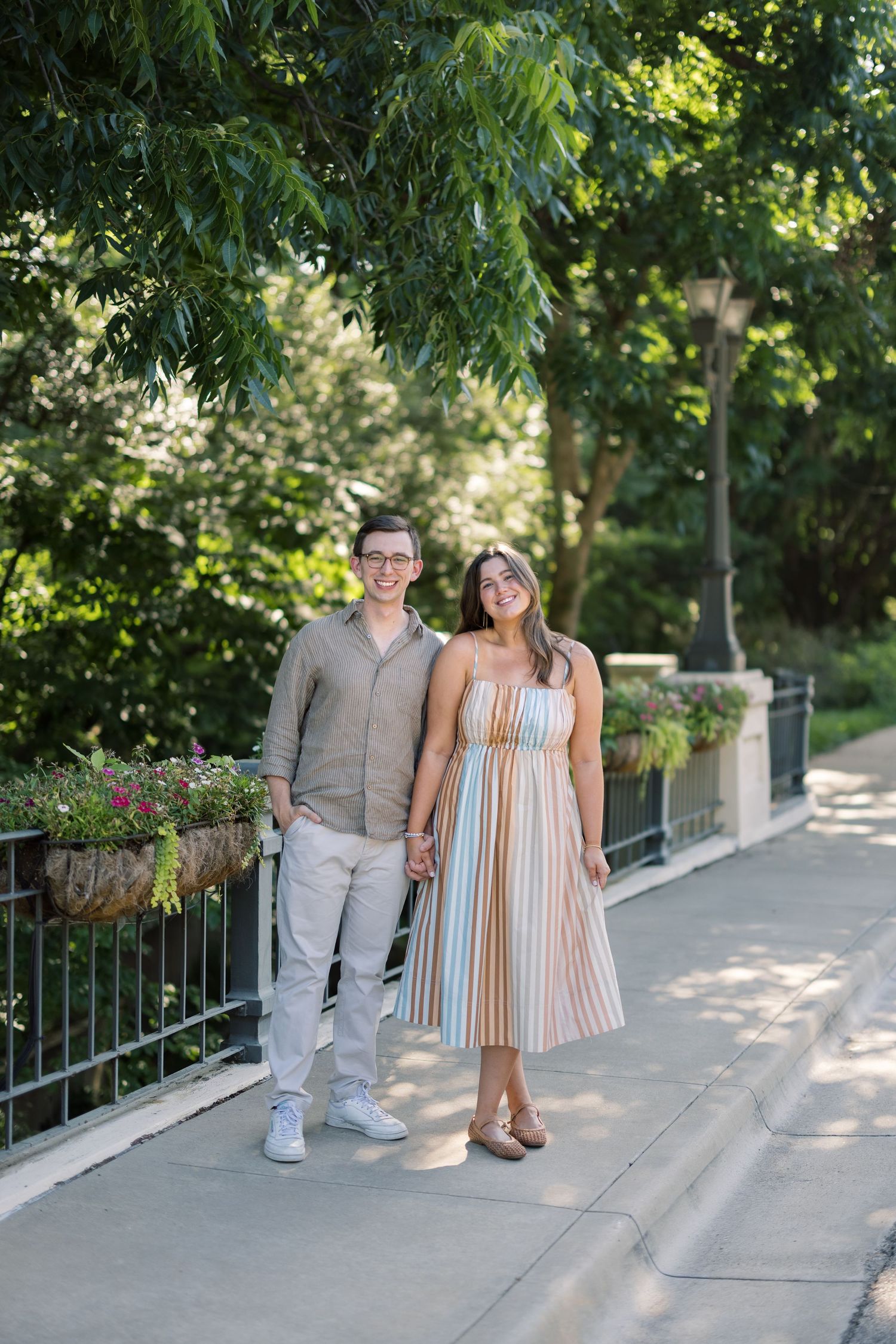 A smiling couple holds hands on a sunny park walkway, man in linen shirt and woman in striped midi dress.