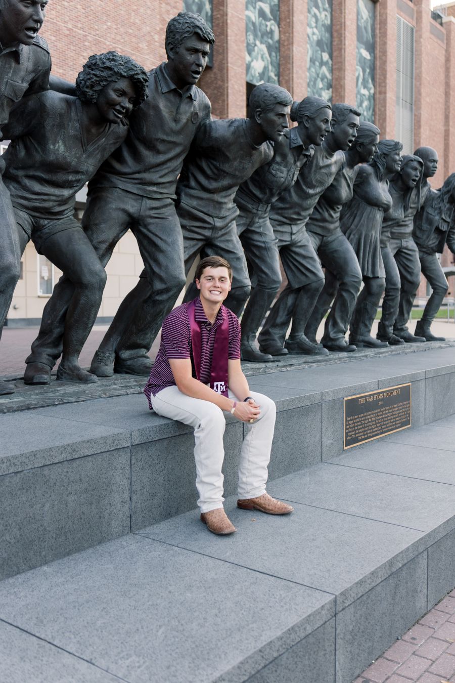Young man in maroon polo sits before a large bronze sculpture of figures at a university campus.
