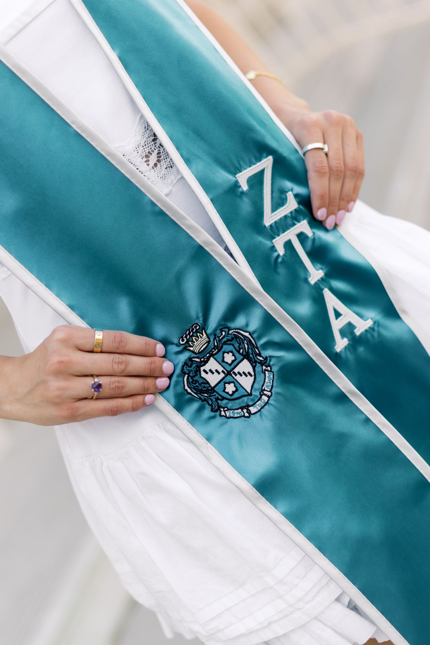 Woman in white dress holding teal ZTA sorority graduation stole with embroidered crest.