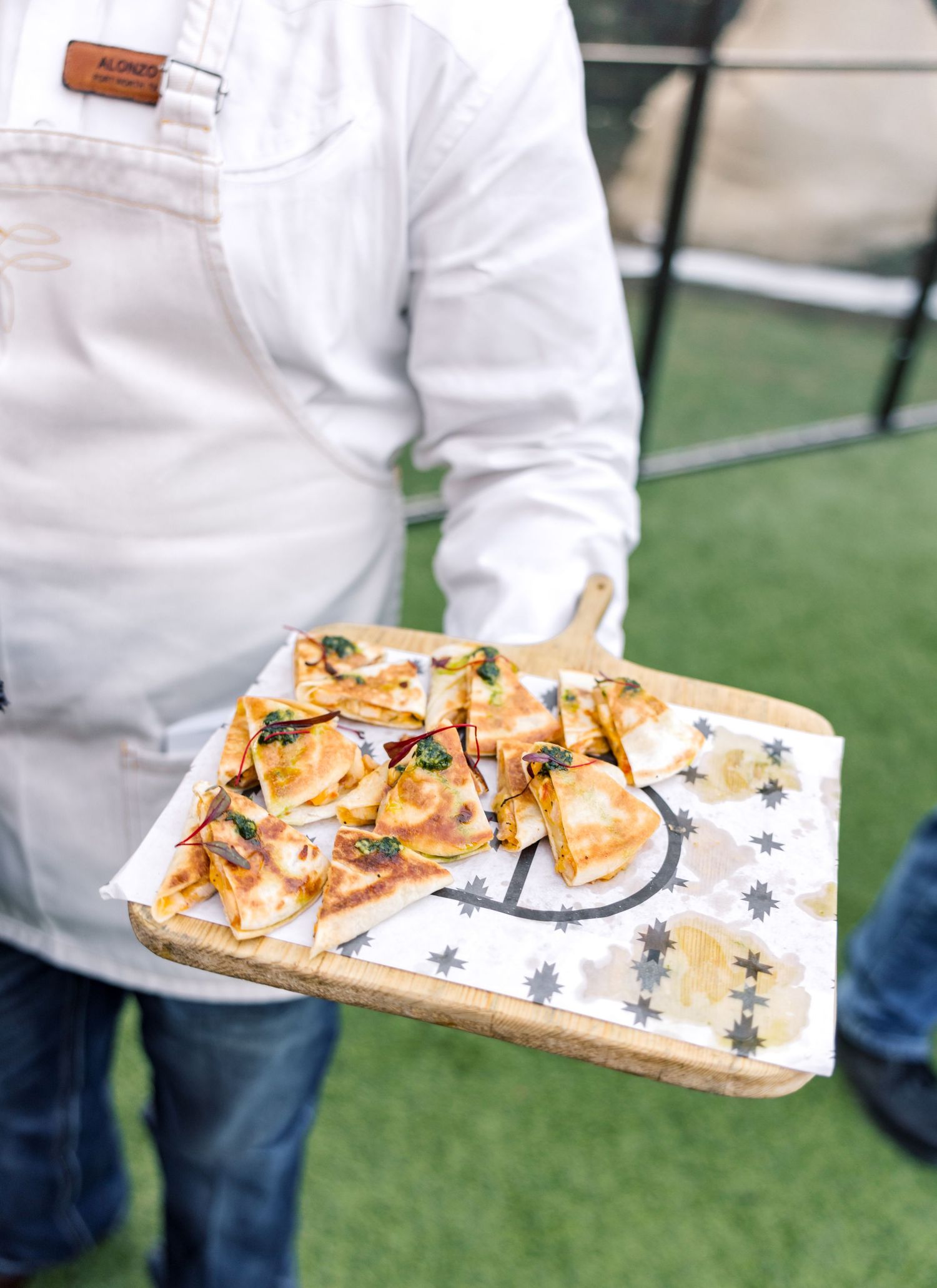 A chef in a white apron holds a wooden board with sliced flatbread pizza topped with cheese and fresh herbs.