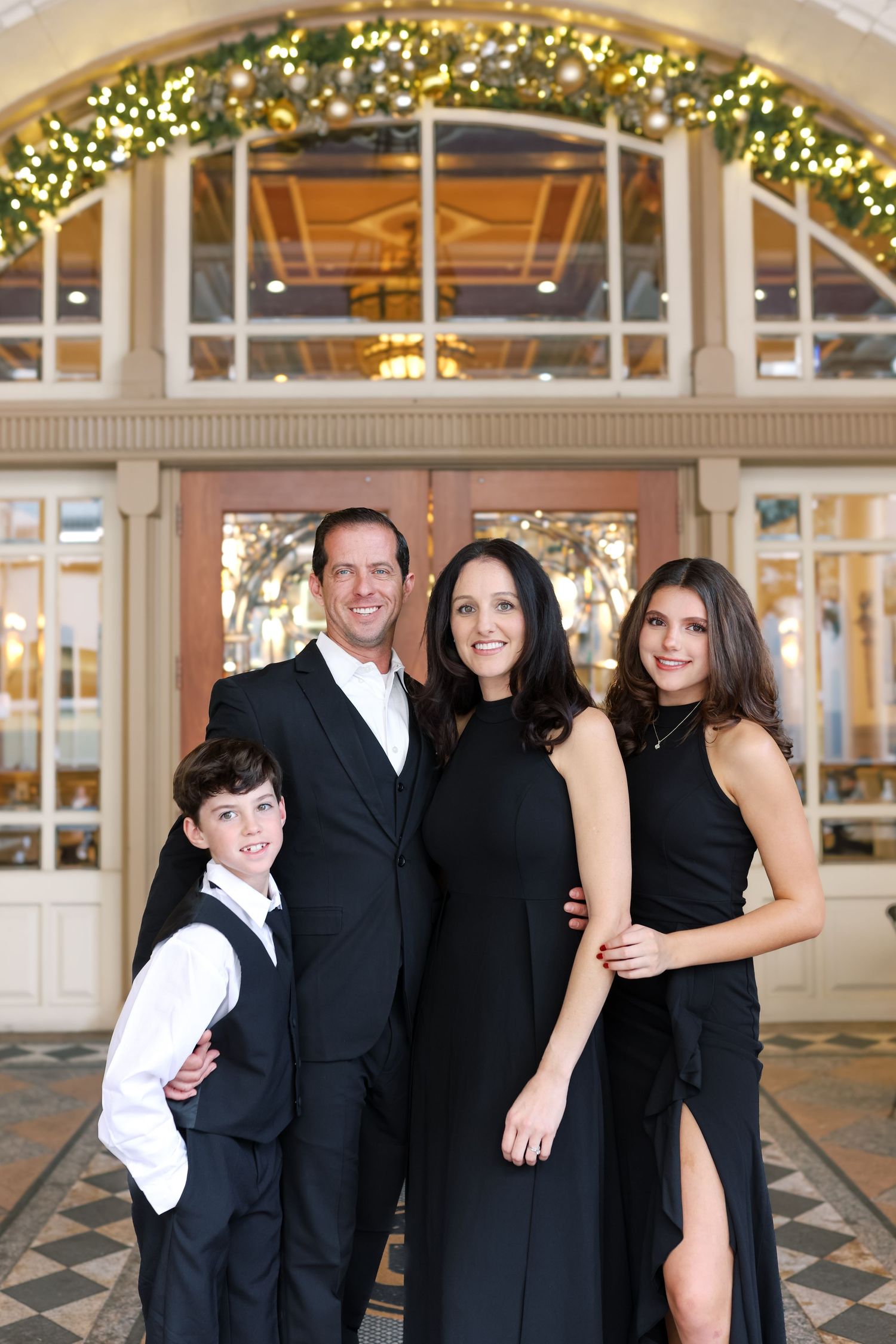 Elegant family of four posing in formal black attire before a festively lit venue entrance.