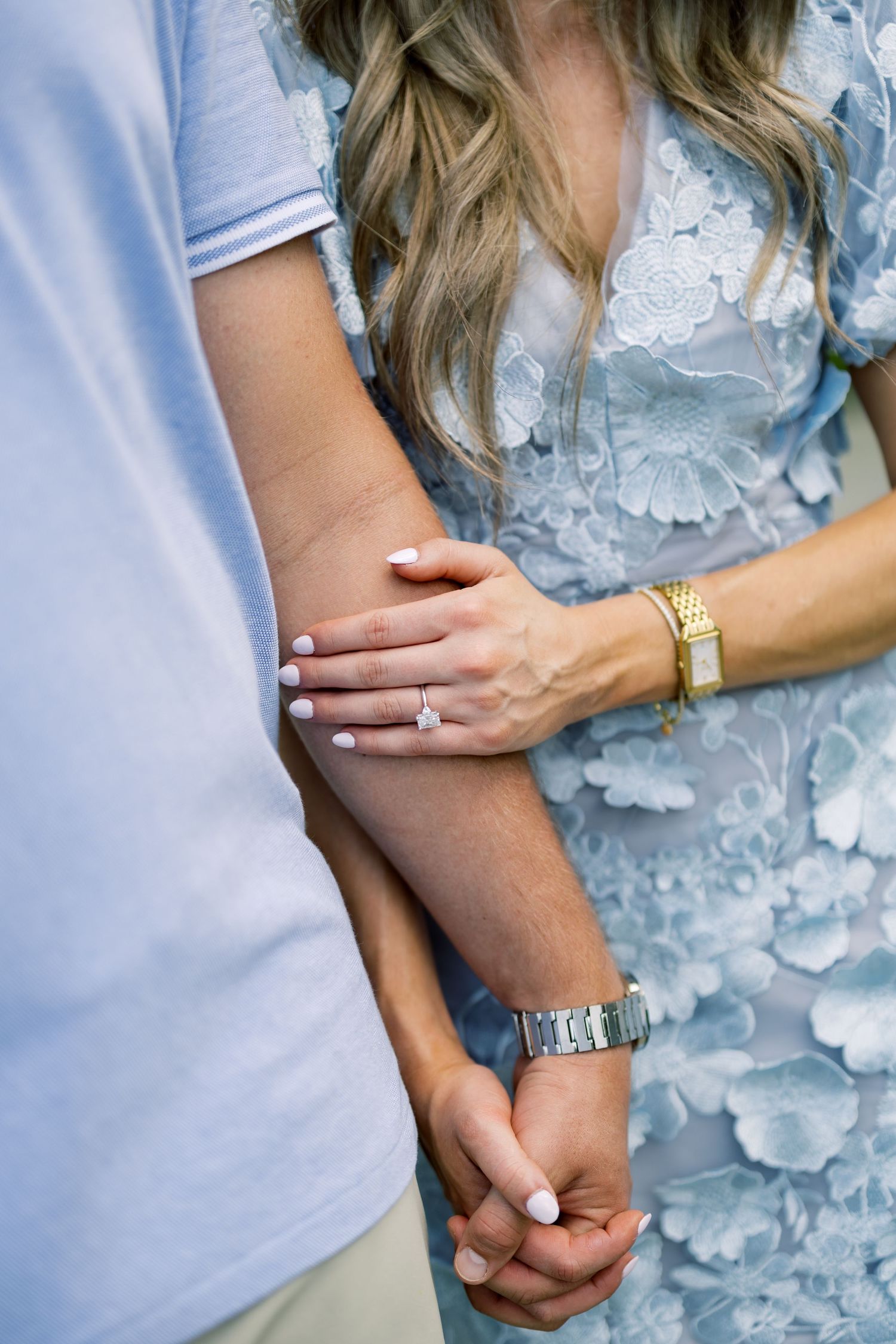 Couple holding hands, woman wearing engagement ring, gold watch, and blue lace dress, man in light blue polo shirt.