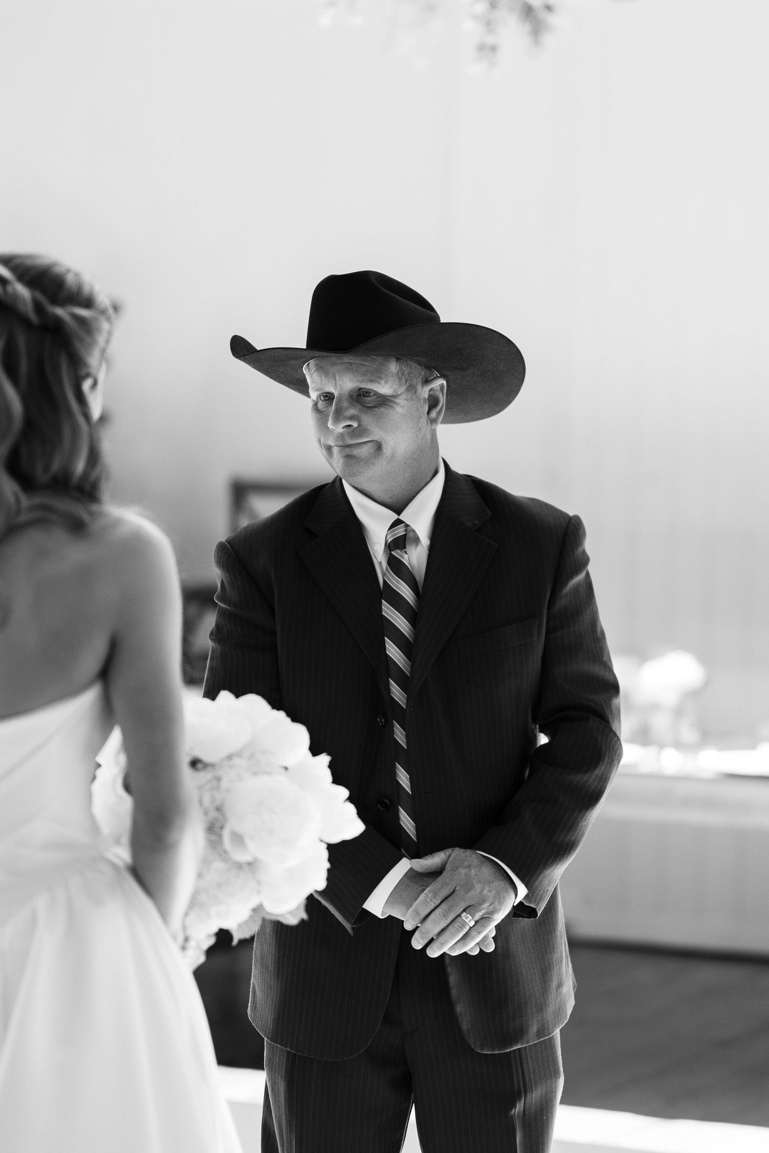 Groom in cowboy hat and suit smiles at bride holding bouquet during outdoor wedding ceremony, black and white photo.