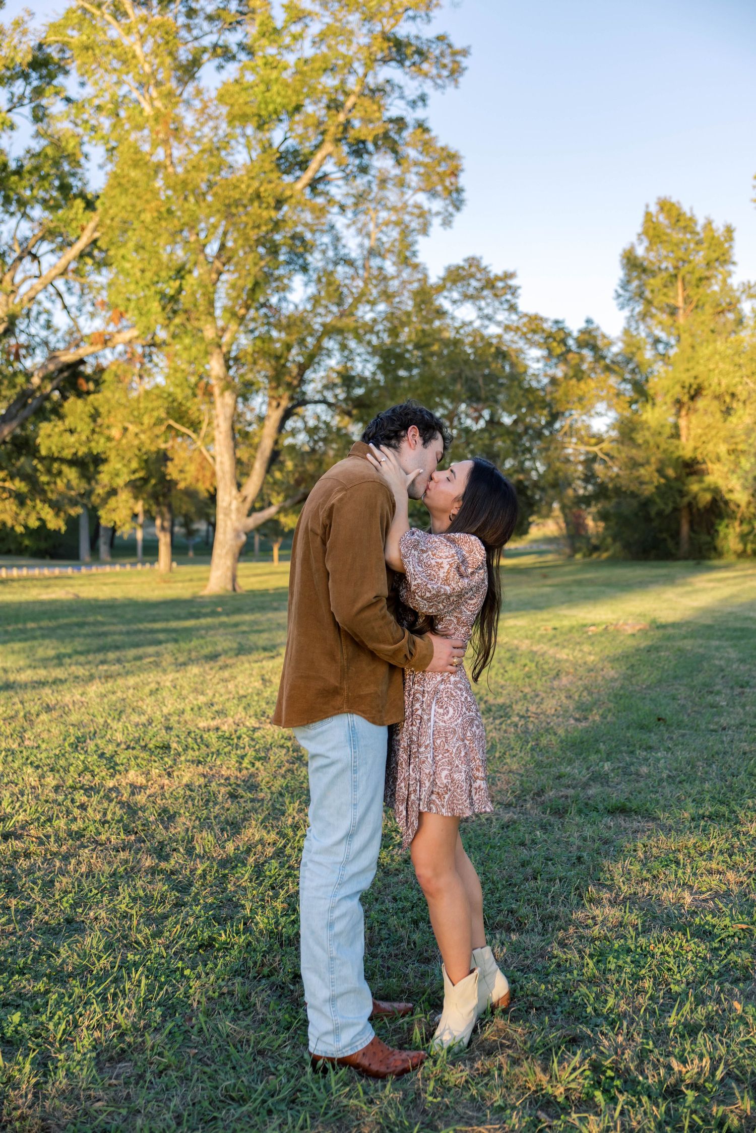 Couple sharing a romantic kiss in a sunlit green park surrounded by tall trees.