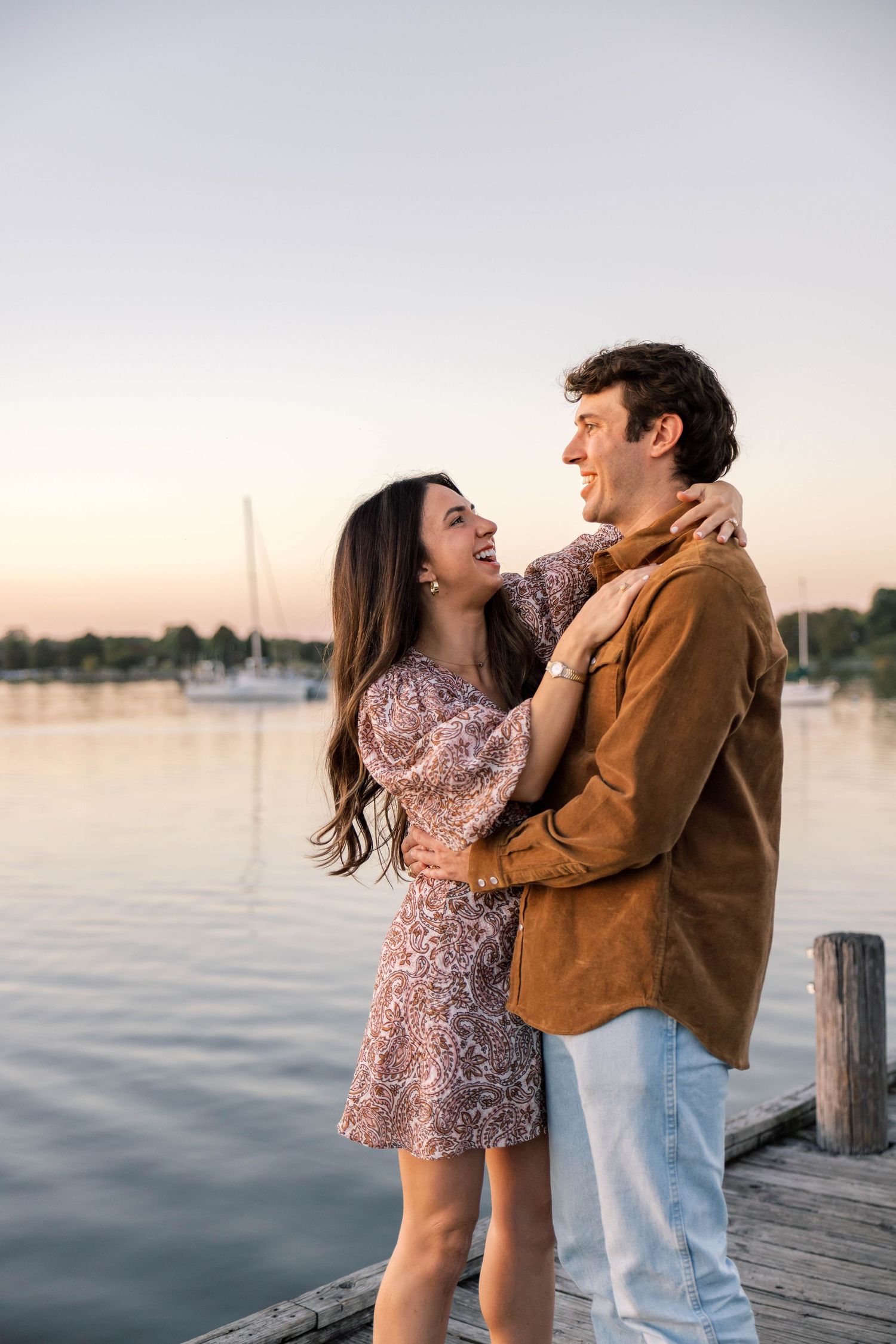 Couple smiling and embracing on a wooden dock at sunset, surrounded by a tranquil lake and sailboats.