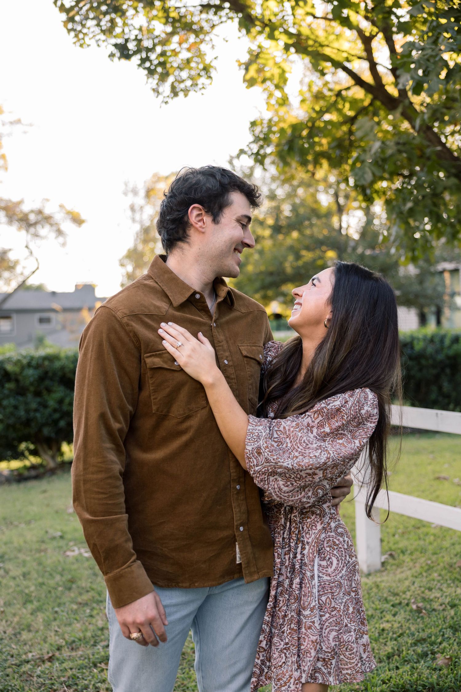 Couple laughing and gazing at each other outdoors, woman in floral dress with hand on mans brown shirt.