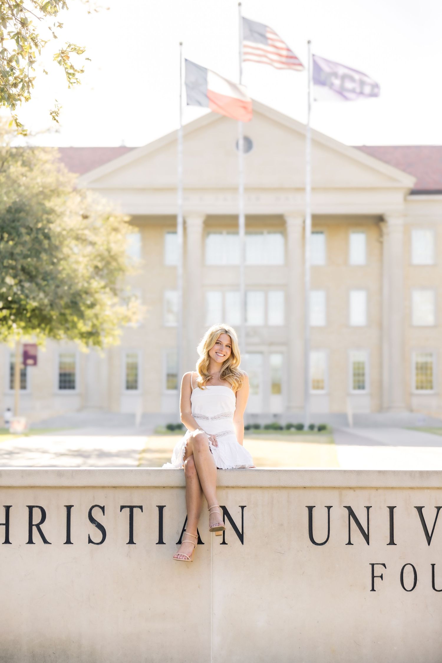 Young woman in white dress sitting on TCU Christian University Foundation wall with flags behind her.