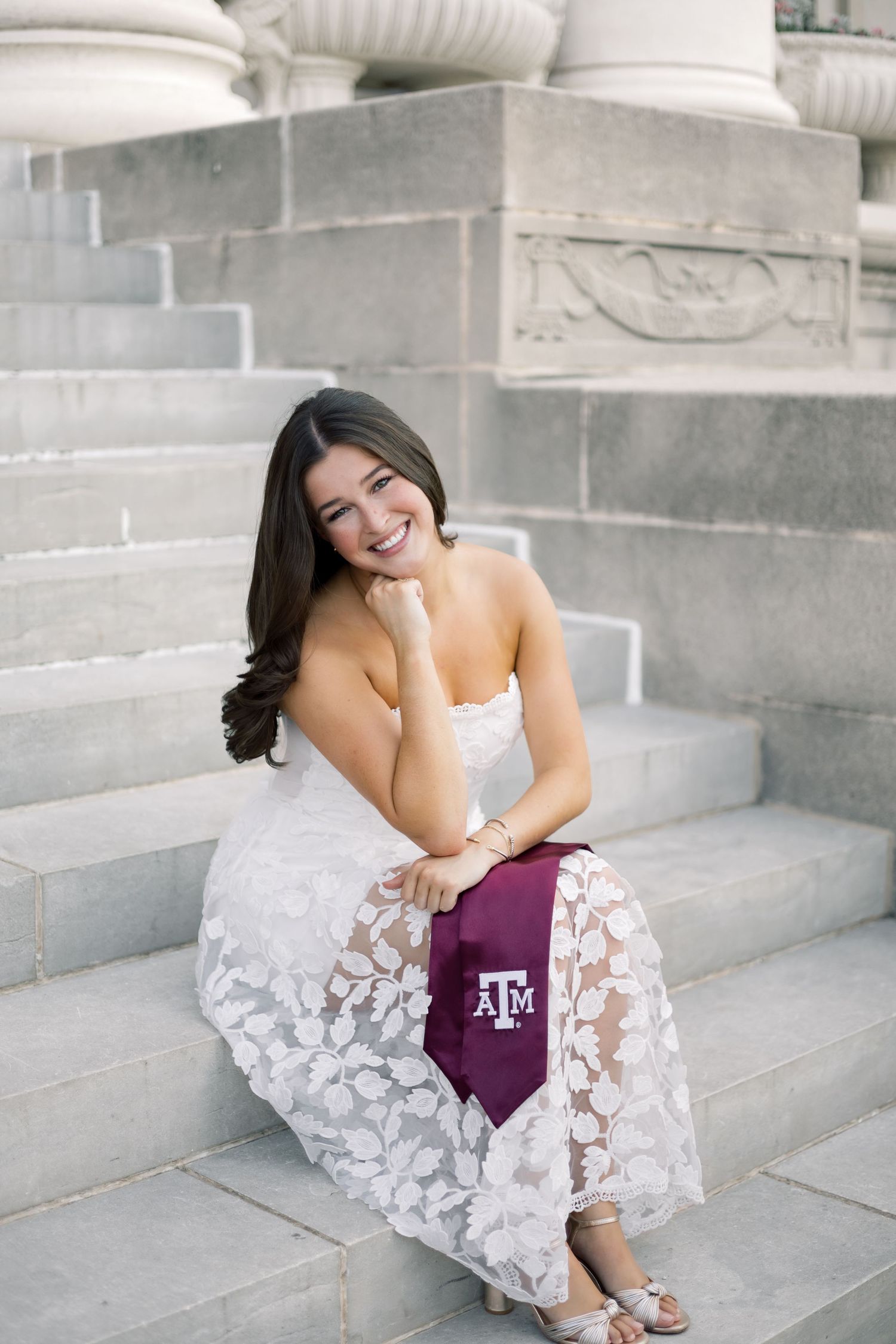 Smiling brunette in strapless white lace dress holding Texas A&M maroon stole on steps.