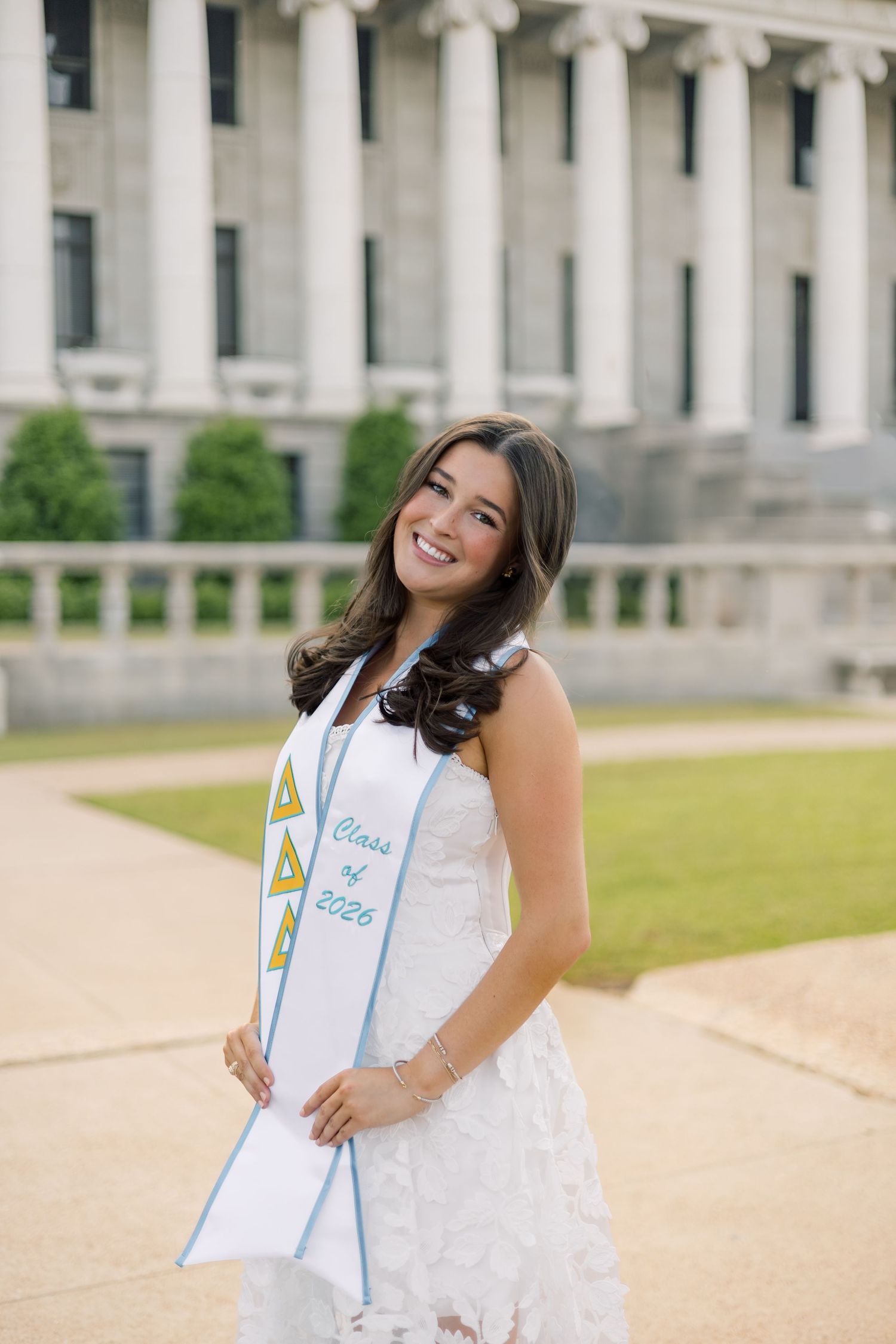 Graduate in white lace dress holding Delta Delta Delta Class of 2026 stole in front of columned building.