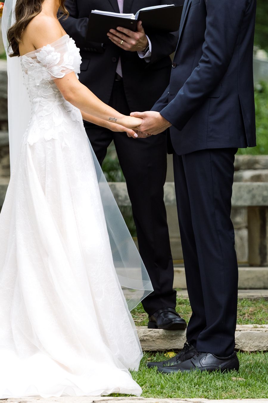 Bride and groom holding hands during outdoor wedding ceremony, officiant holding book in background.
