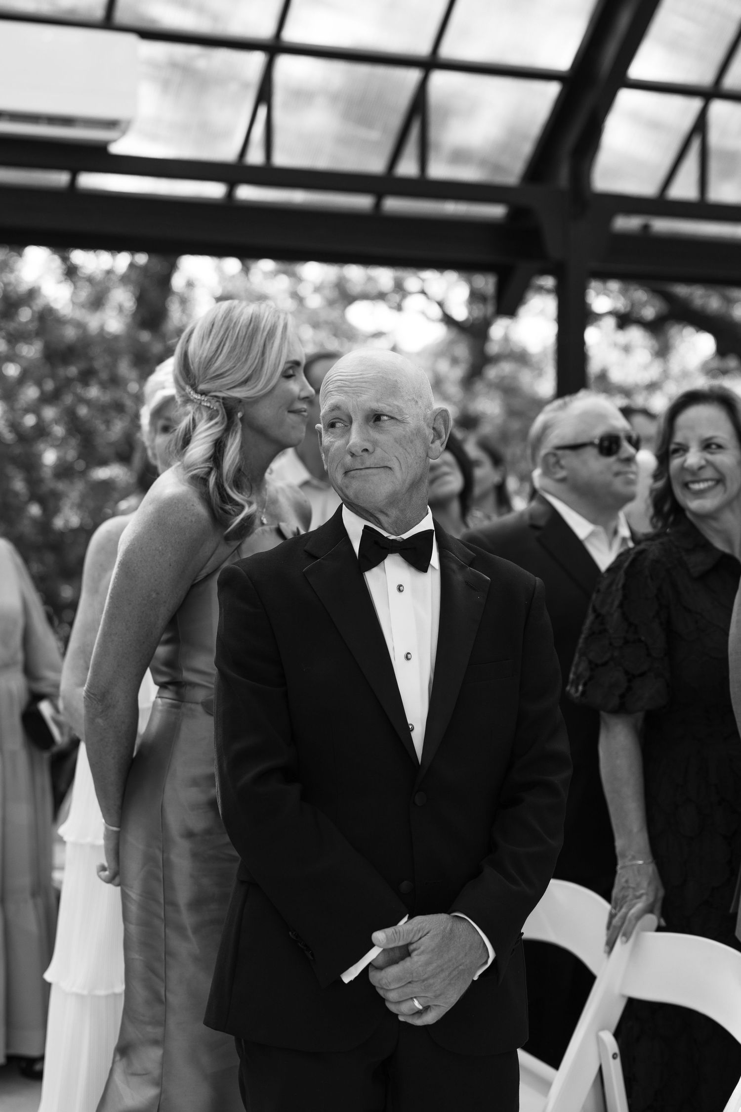 Black and white photo of elegantly dressed wedding guests, man in tuxedo and woman in gown, at an outdoor ceremony.