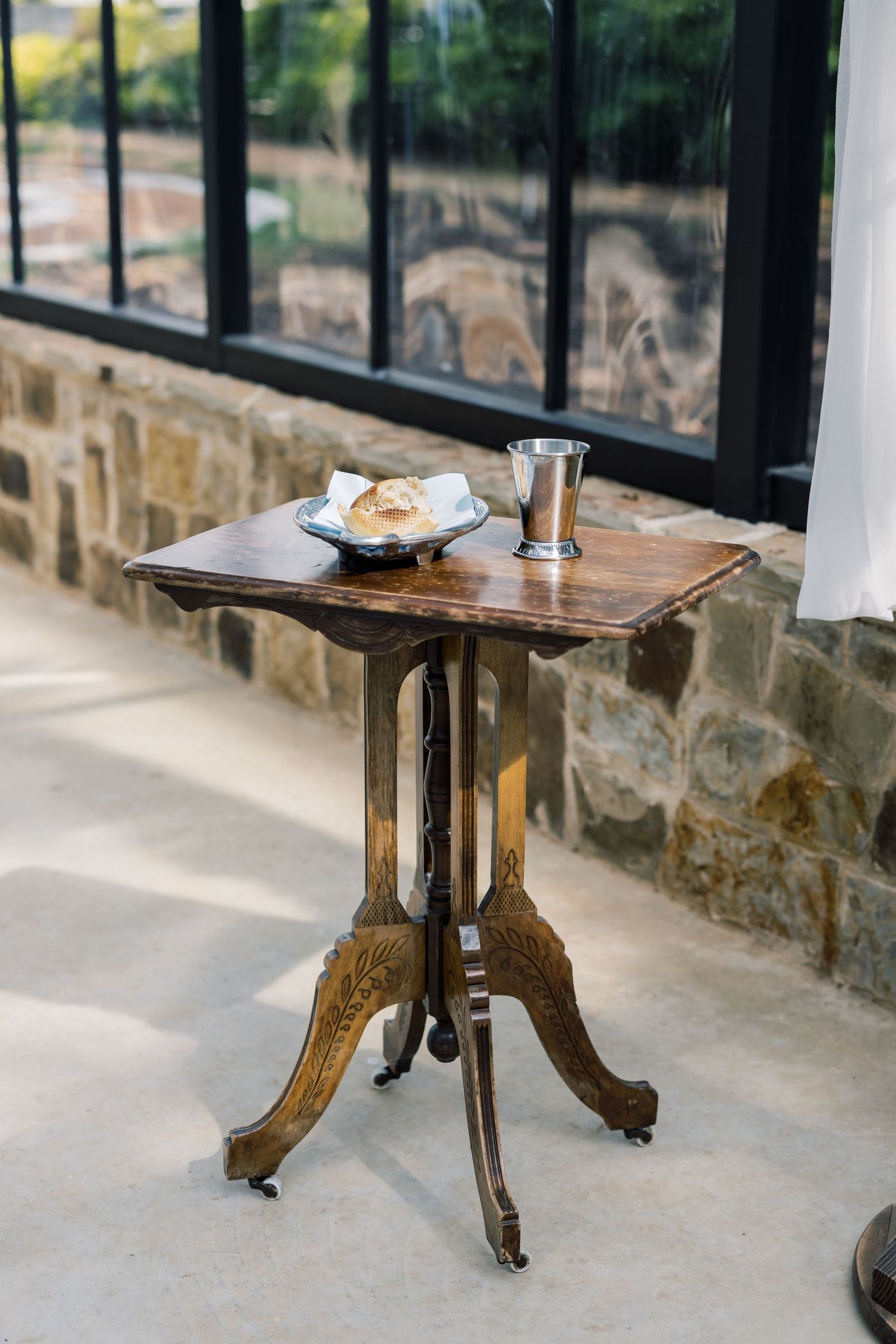 Antique wooden side table with ornate carved legs on casters, holding a silver cup and bread plate near a stone wall window.