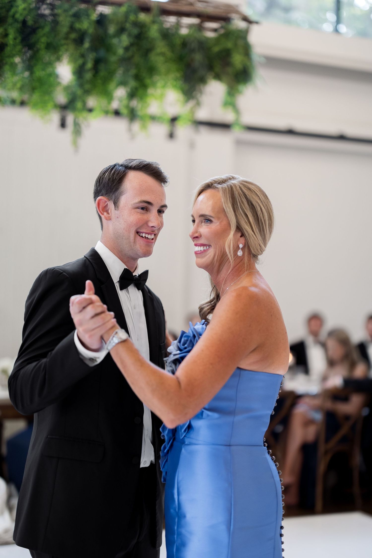 Groom in black tuxedo and woman in blue strapless gown share a joyful mother-son dance at elegant wedding reception.