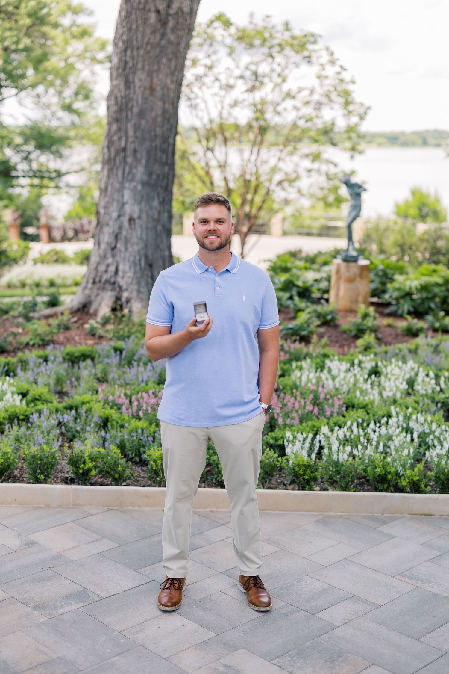 Man in light blue polo holds open ring box in a garden setting with lush greenery and a water view.
