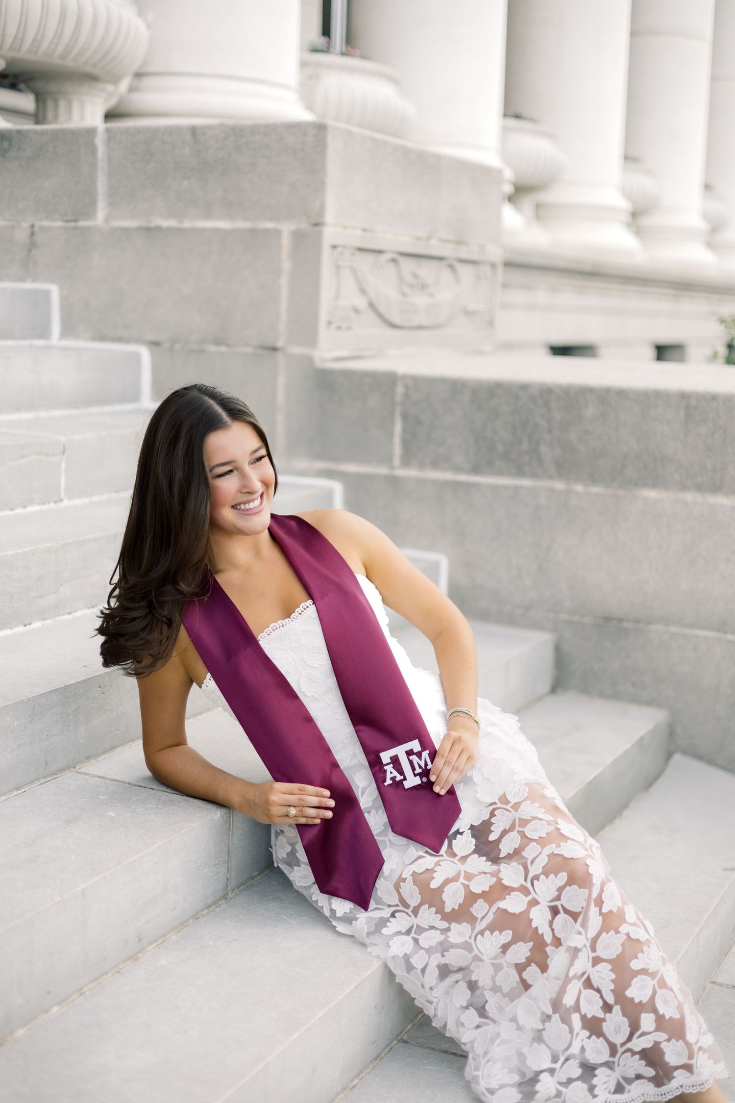 Smiling woman in white lace dress with maroon Texas A&M graduation stole sitting on marble steps.