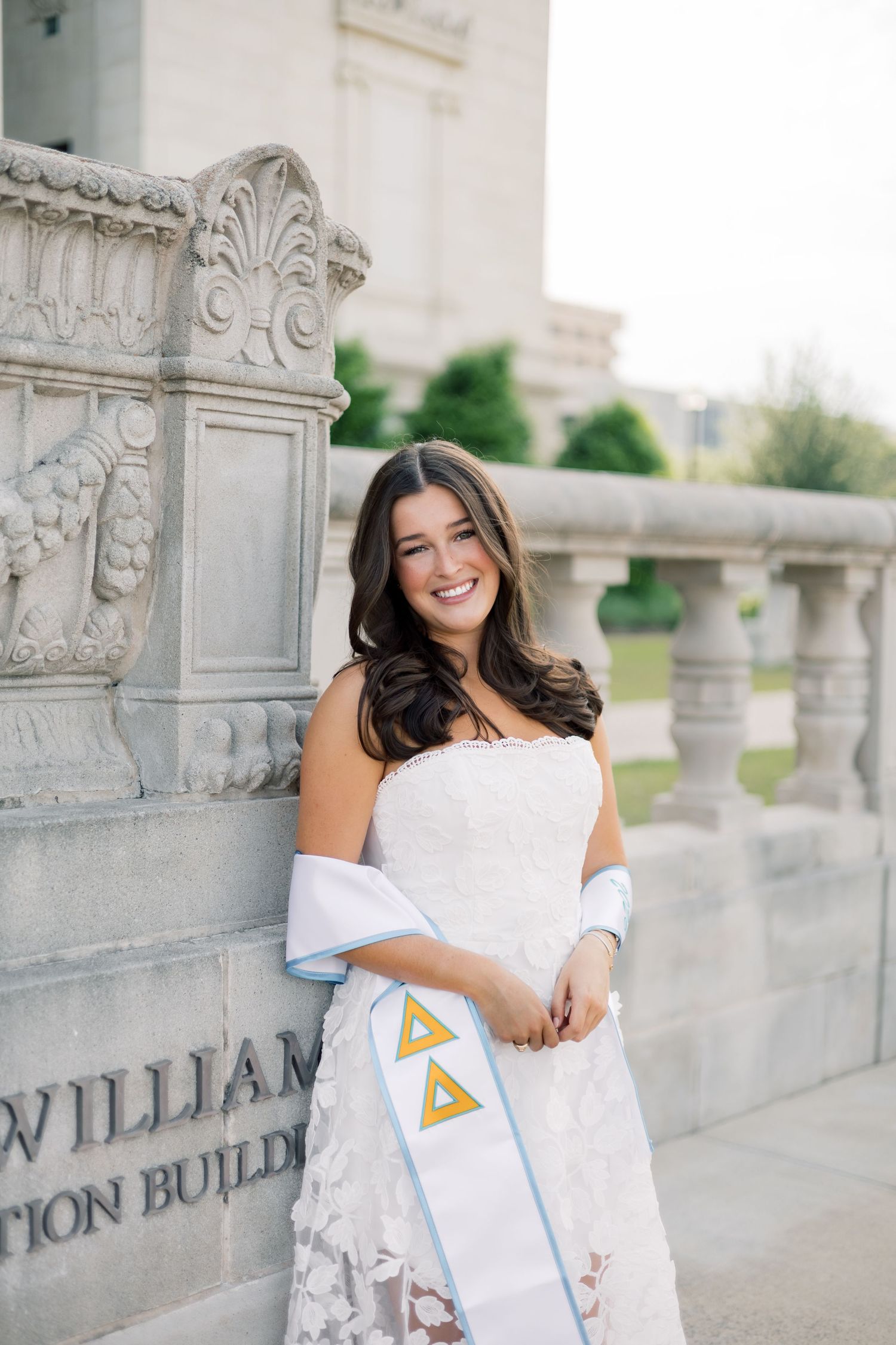 Smiling brunette graduate in white strapless dress with Delta Delta Delta stole at Texas A&M campus.