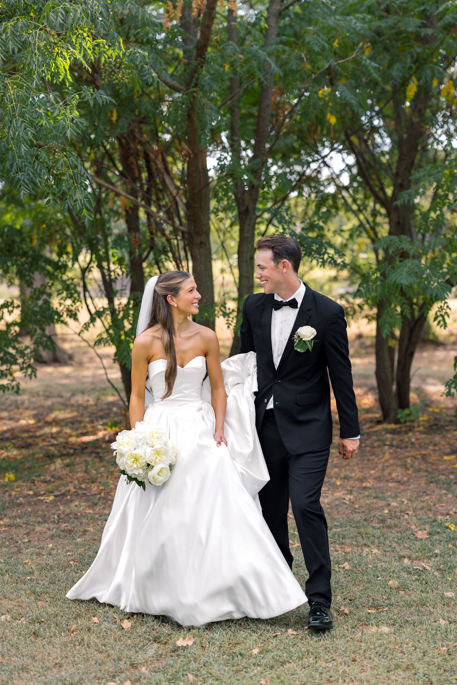 A bride and groom walk hand-in-hand through a lush green garden, smiling at each other on their wedding day.