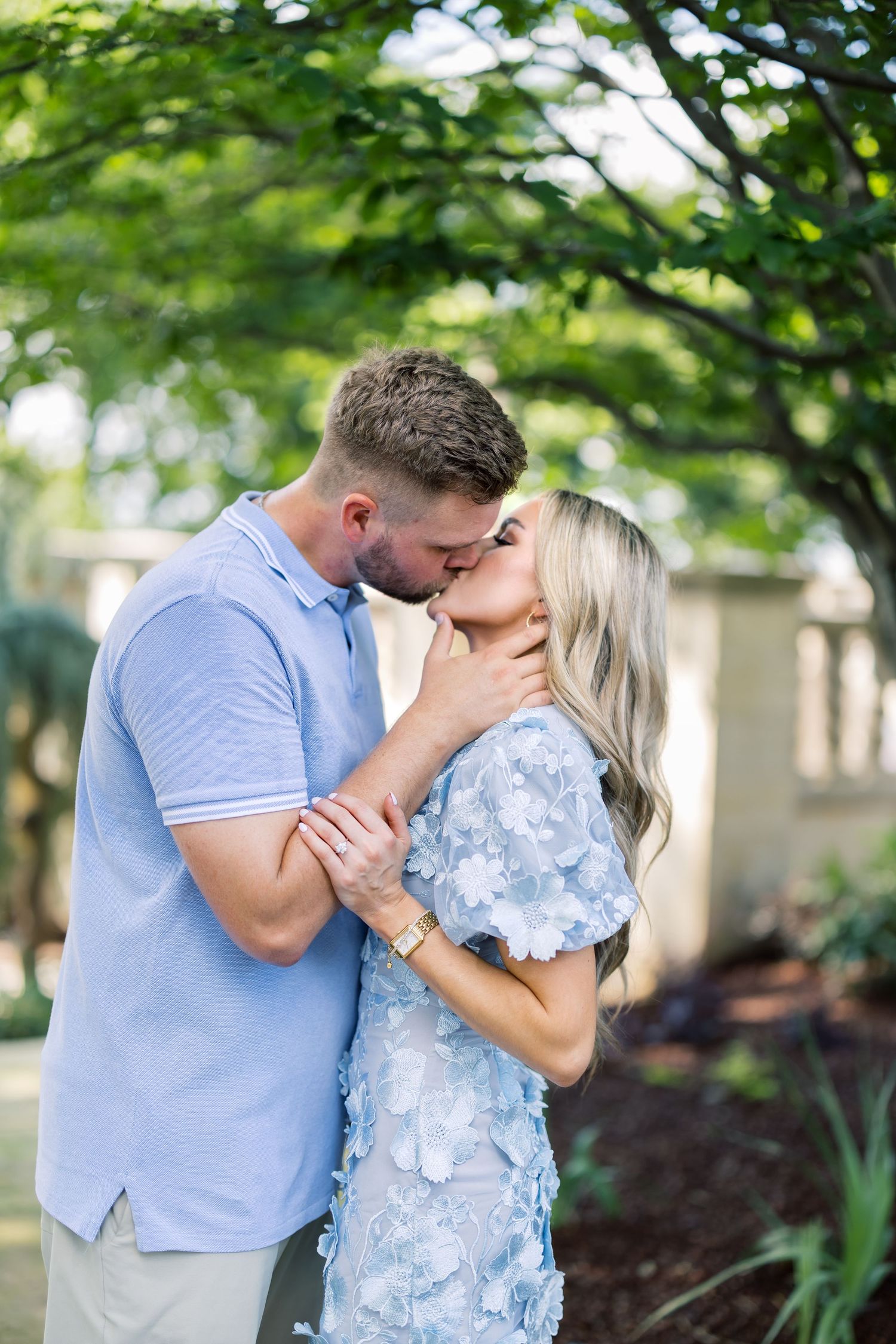 A couple shares a romantic kiss outdoors, dressed in coordinating blue outfits, surrounded by lush green trees.