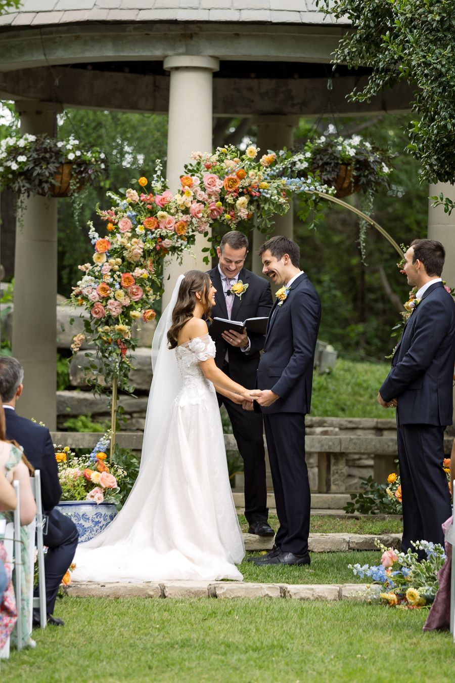 Bride and groom exchange vows under floral arch at outdoor gazebo wedding ceremony with colorful blooms.