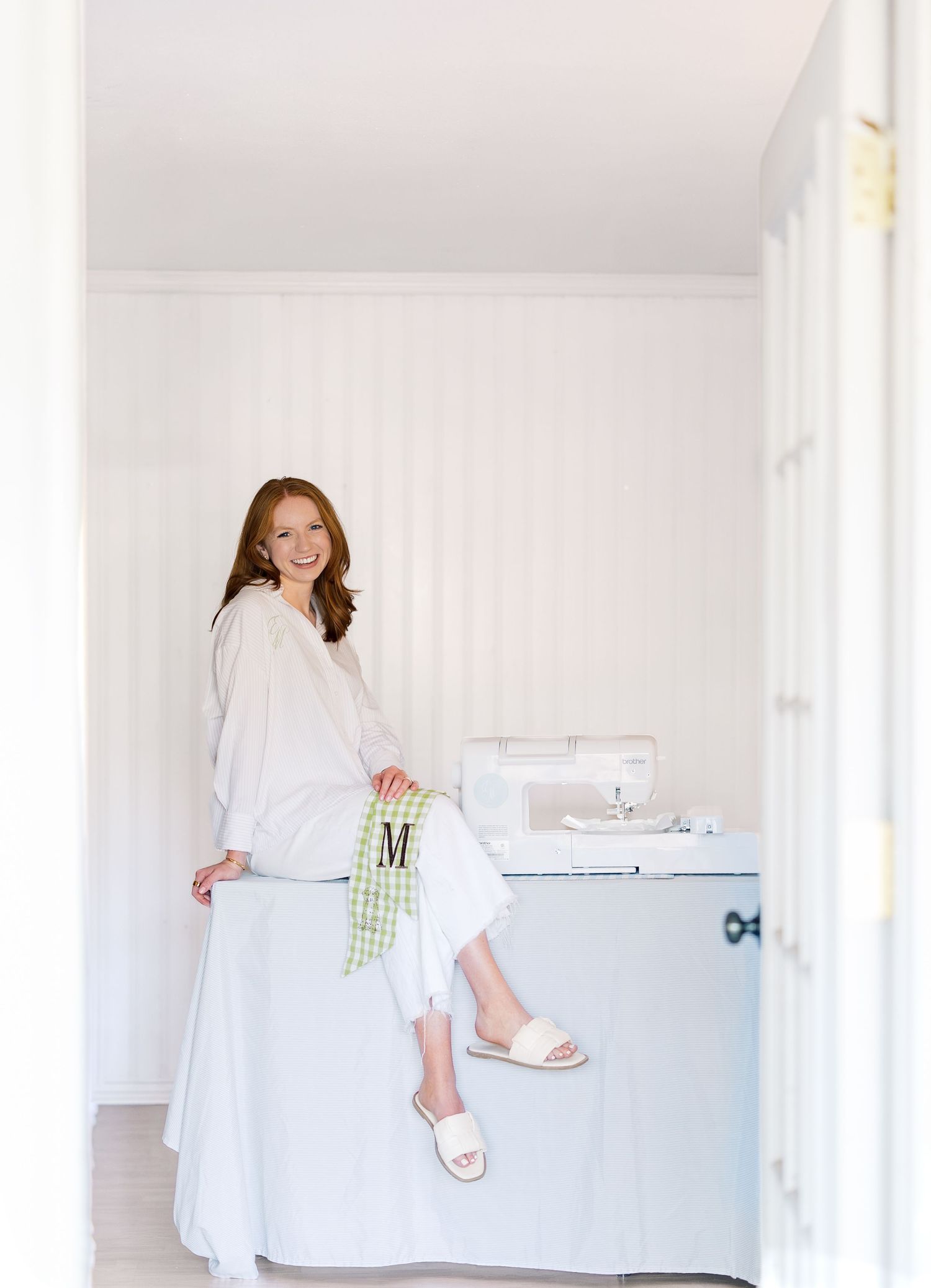 Smiling woman in white outfit sitting on a light blue cabinet in a bright white room with a sewing machine.