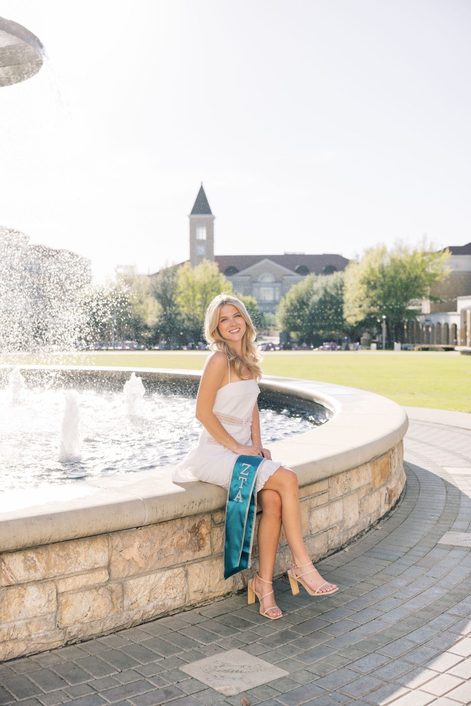 Smiling graduate in white dress with teal sash seated on stone fountain on sunny campus.