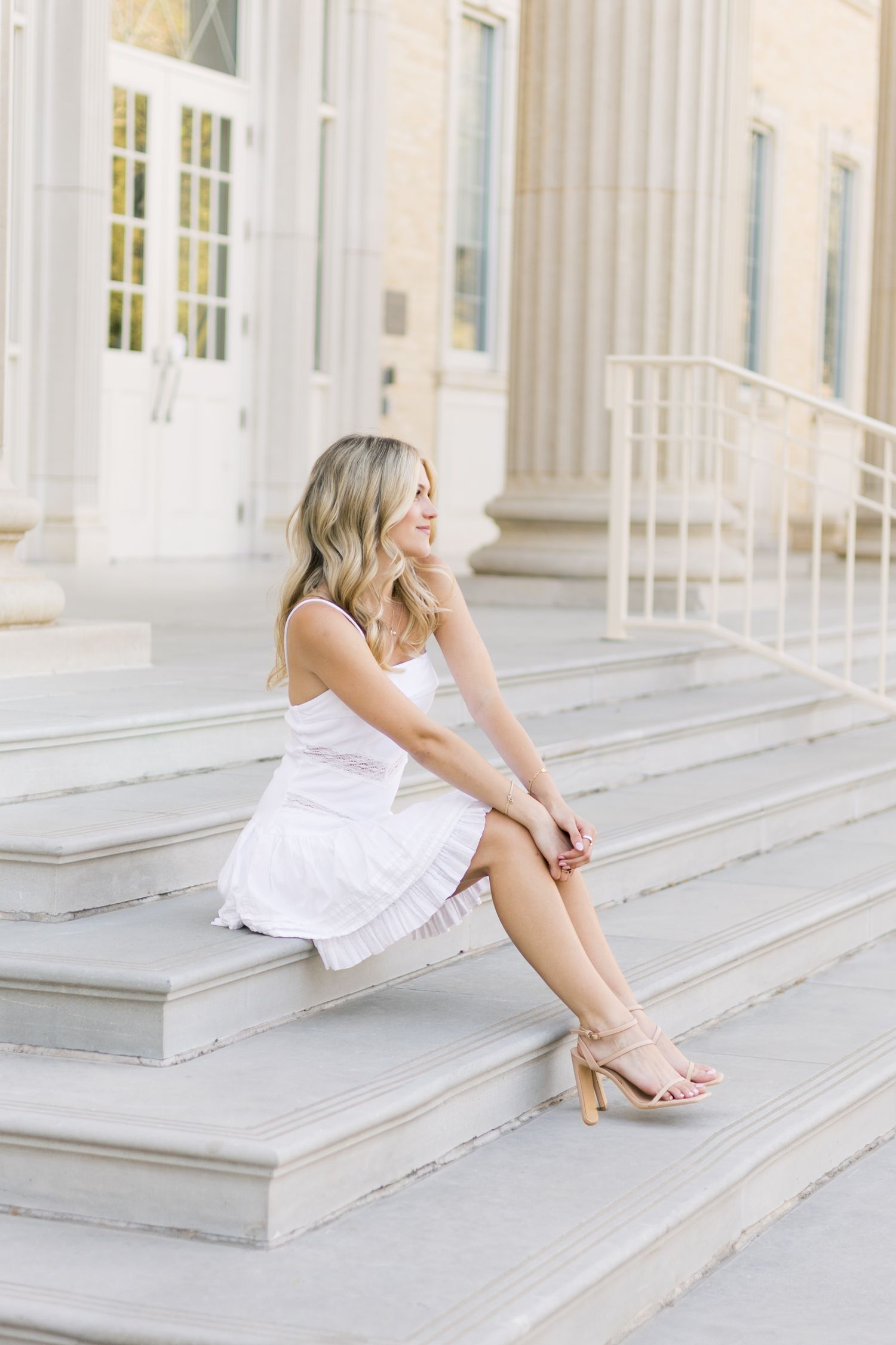 Blonde woman in white sundress sitting on marble steps of a neoclassical building, looking away.