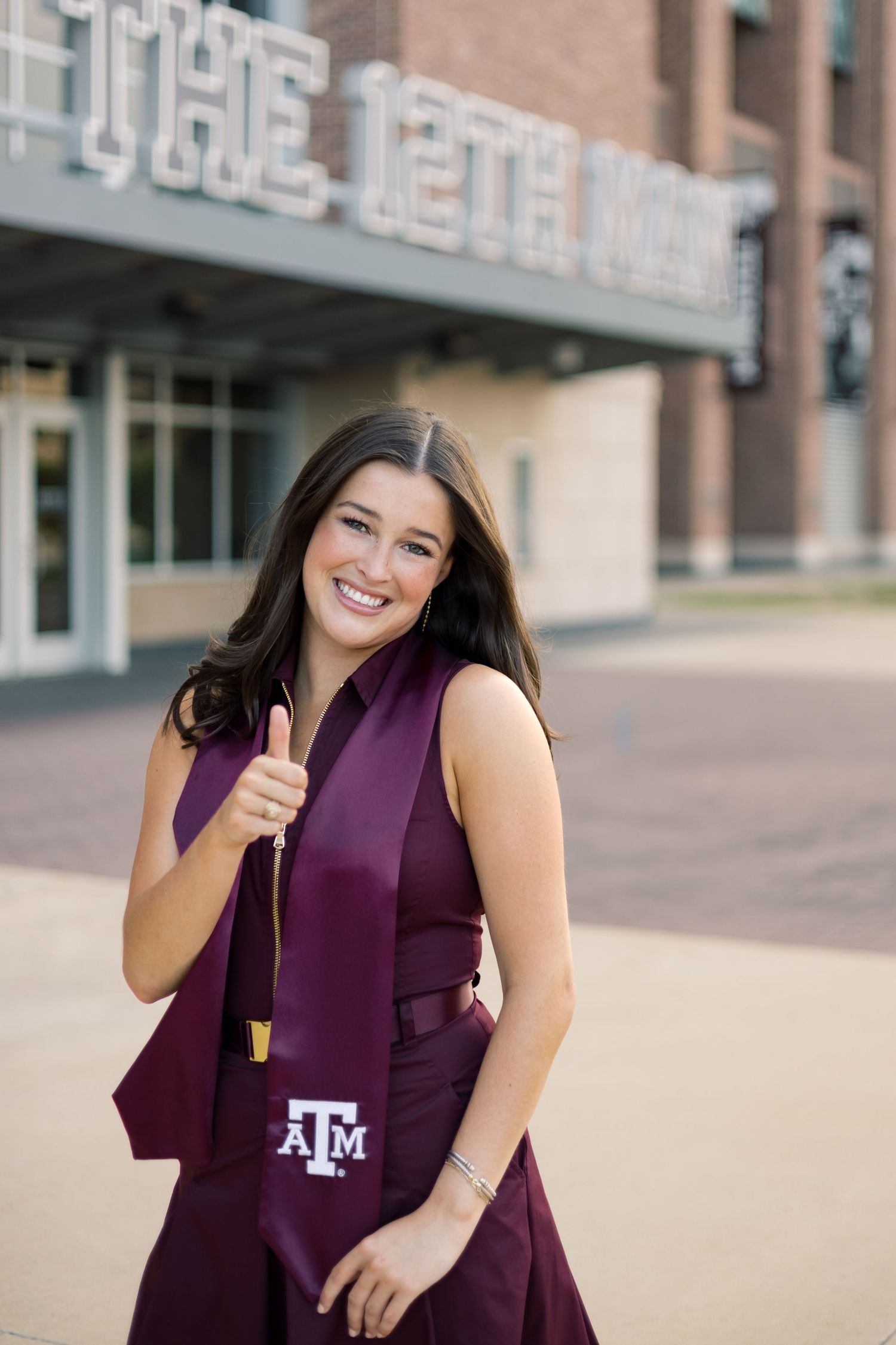 Happy Texas A&M graduate in maroon dress and stole giving thumbs up at The 12th Man stadium.
