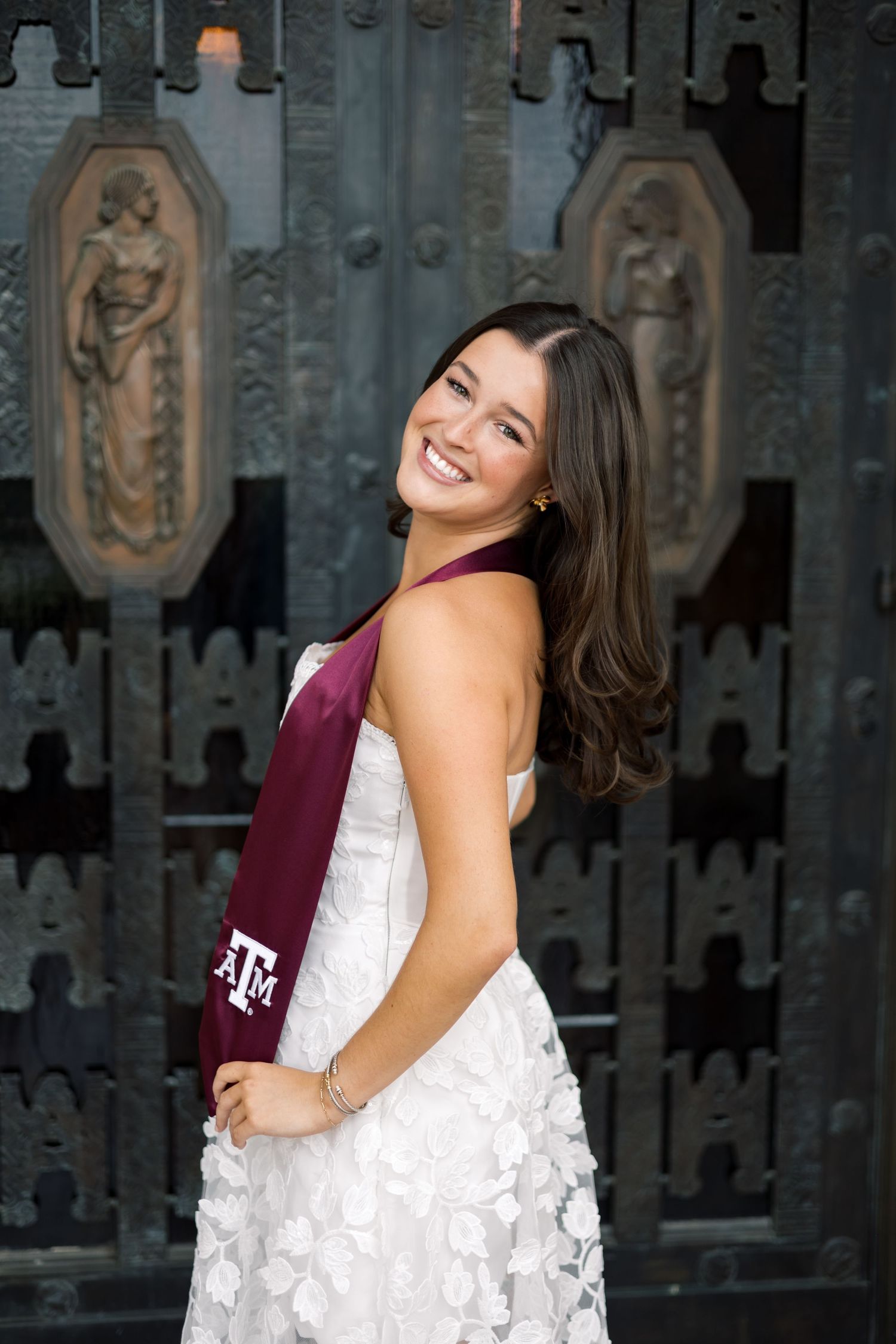 Smiling Texas A&M graduate in white lace dress with maroon ATM stole poses before ornate bronze doors.