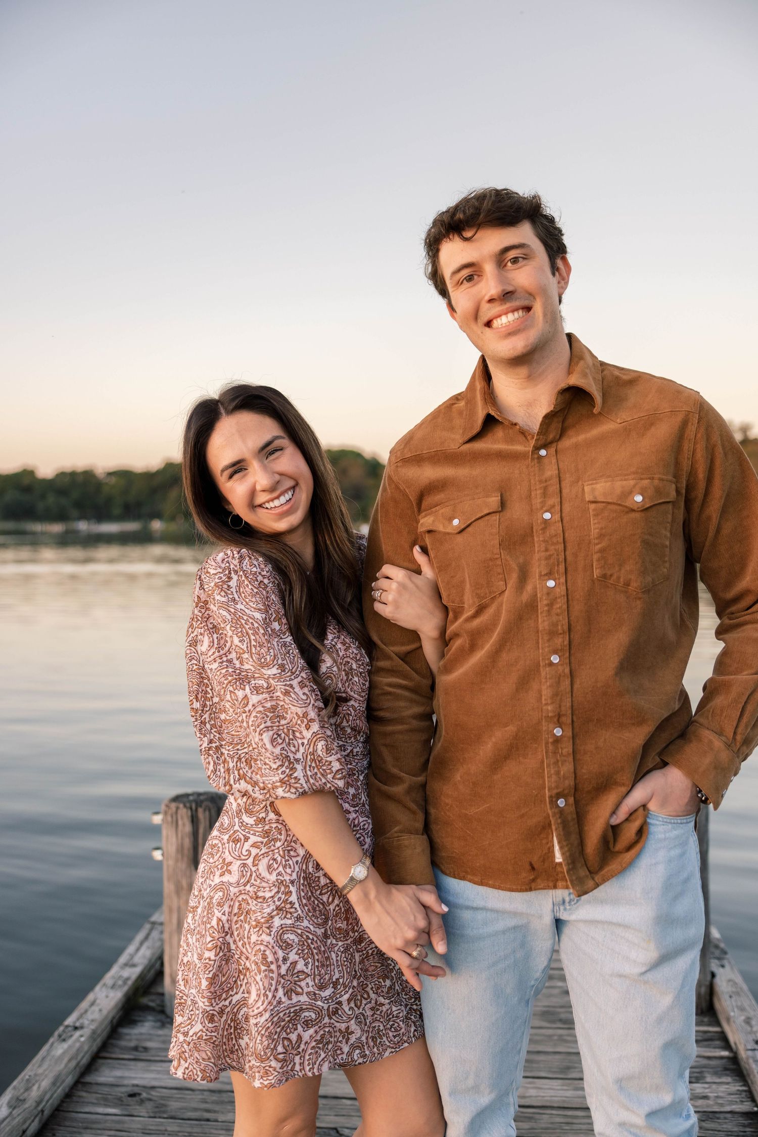 Smiling couple holding hands on a wooden dock with a serene lake and golden sunset behind them.