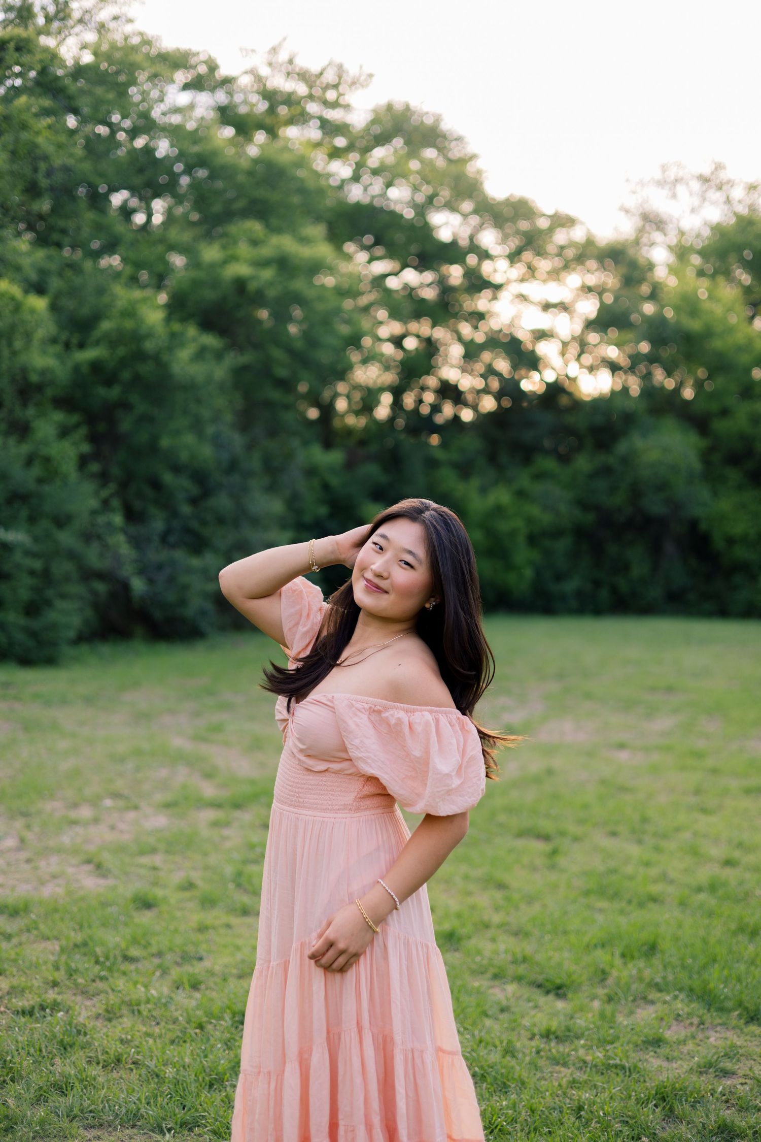 Young woman in pink off-shoulder dress posing in green field at golden hour.
