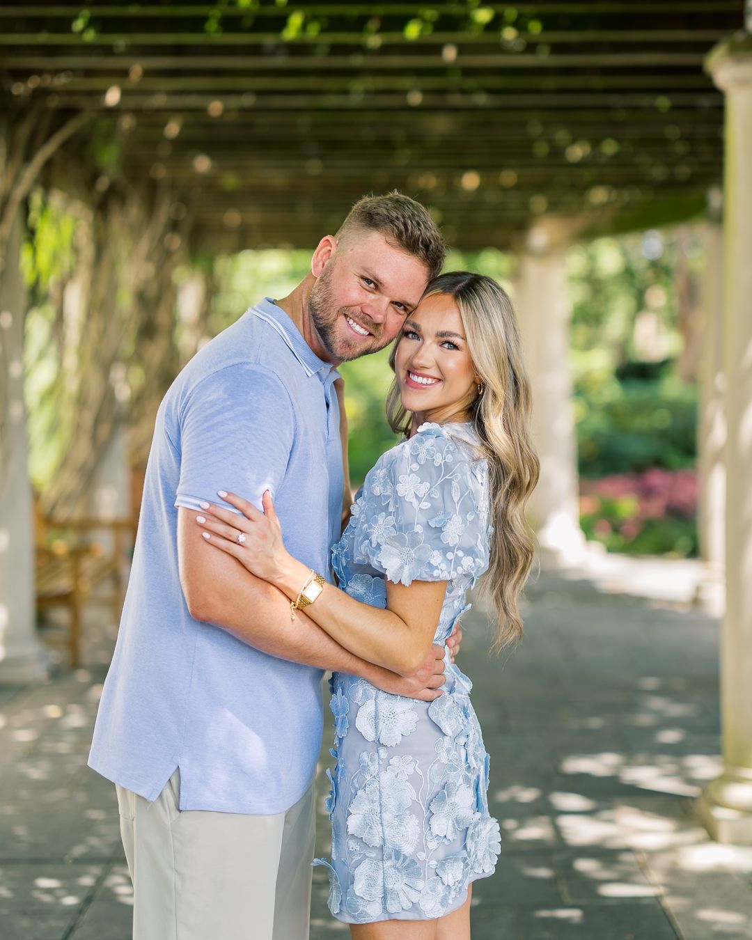 A smiling couple embraces under a garden pergola, wearing coordinating light blue outfits in a romantic outdoor setting.