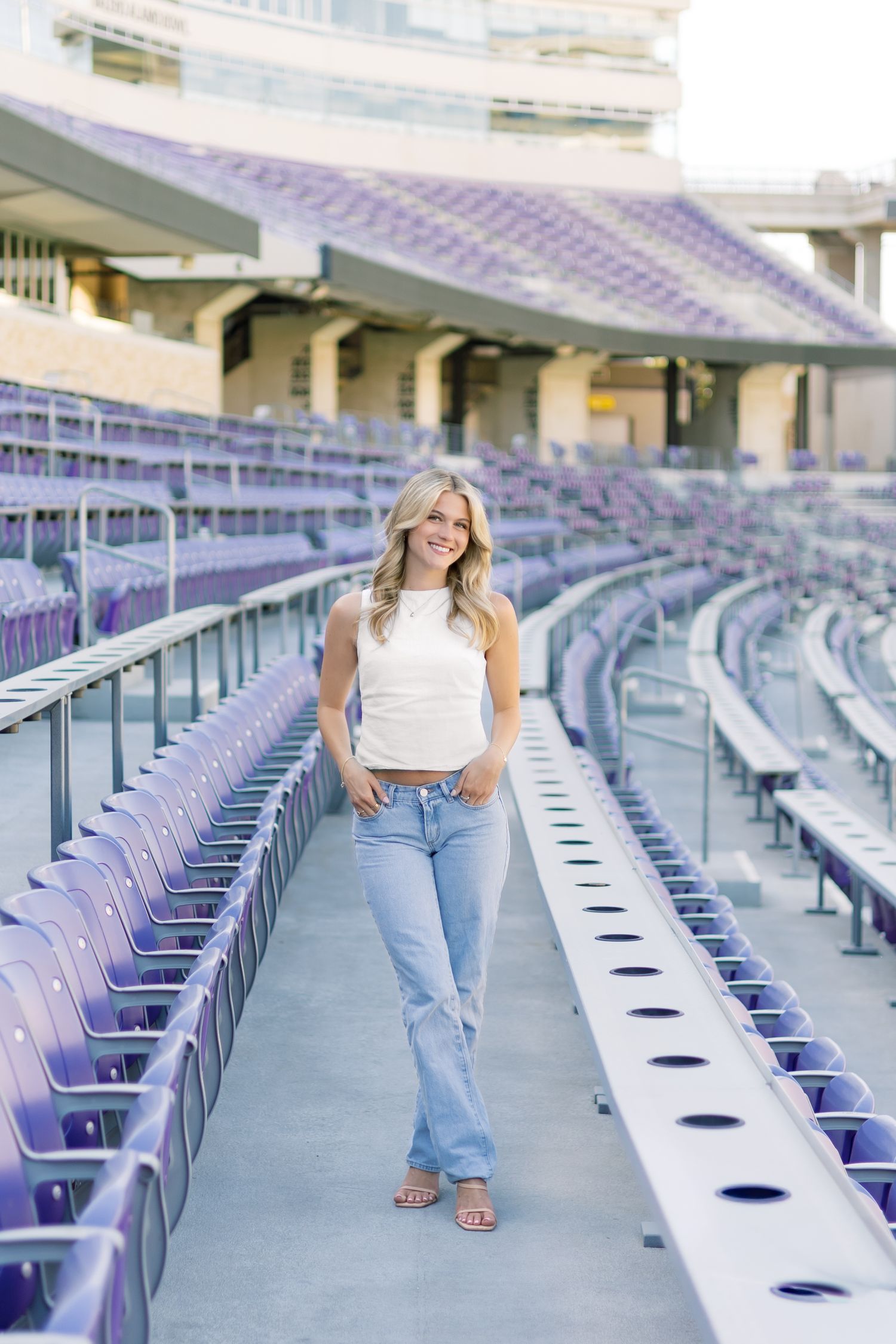 Smiling blonde woman in white crop top and jeans posing in purple TCU stadium bleachers.