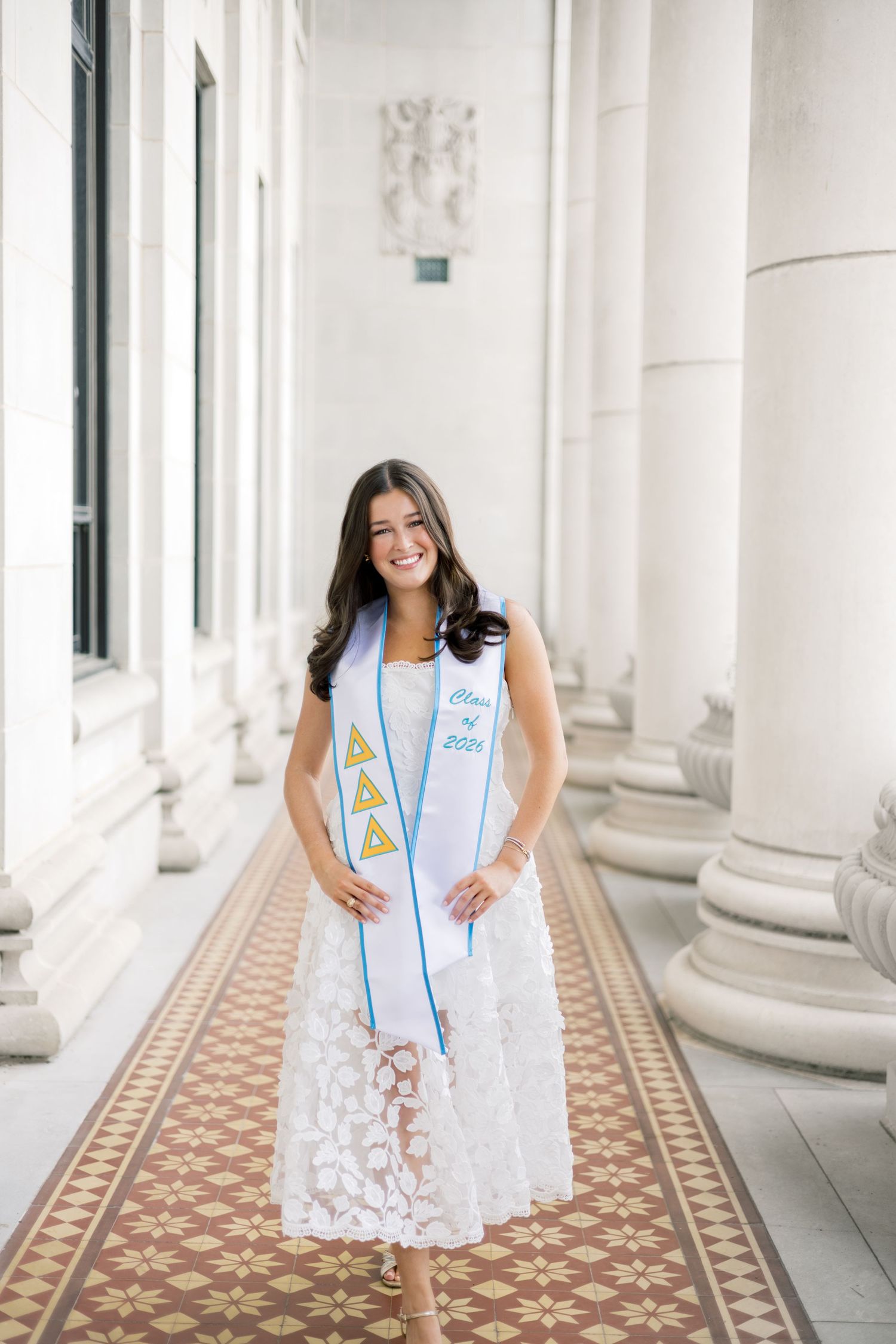 Tri Delta Class of 2024 graduate in white lace dress wears sorority stole in elegant columned hallway.