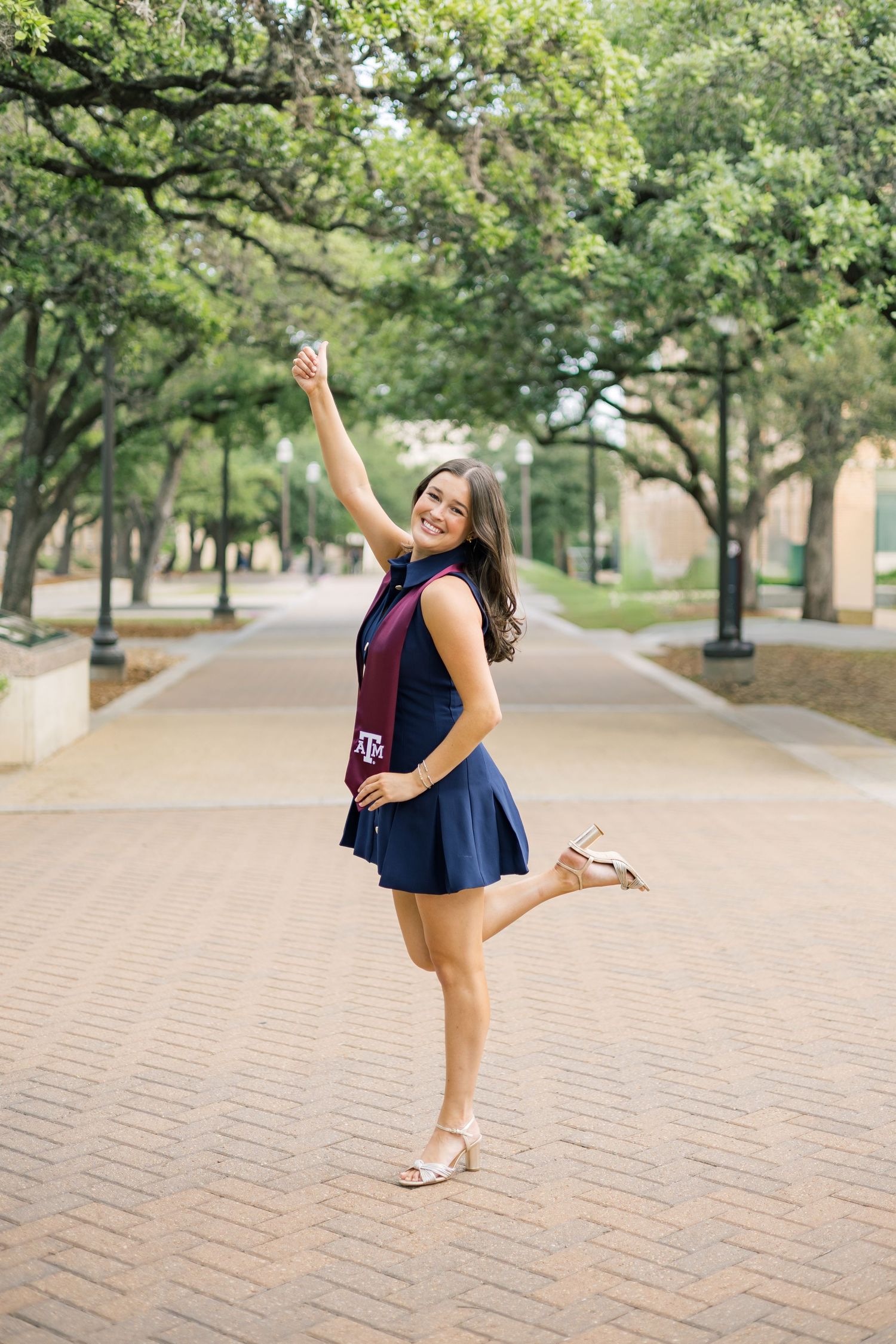 Excited Texas A&M graduate celebrates on brick pathway in navy dress and maroon stole under oak trees.